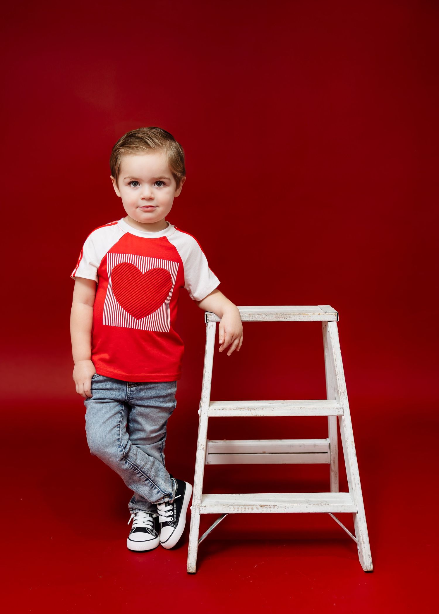 Young boy in red heart raglan shirt and jeans leaning against white ladder on red background.