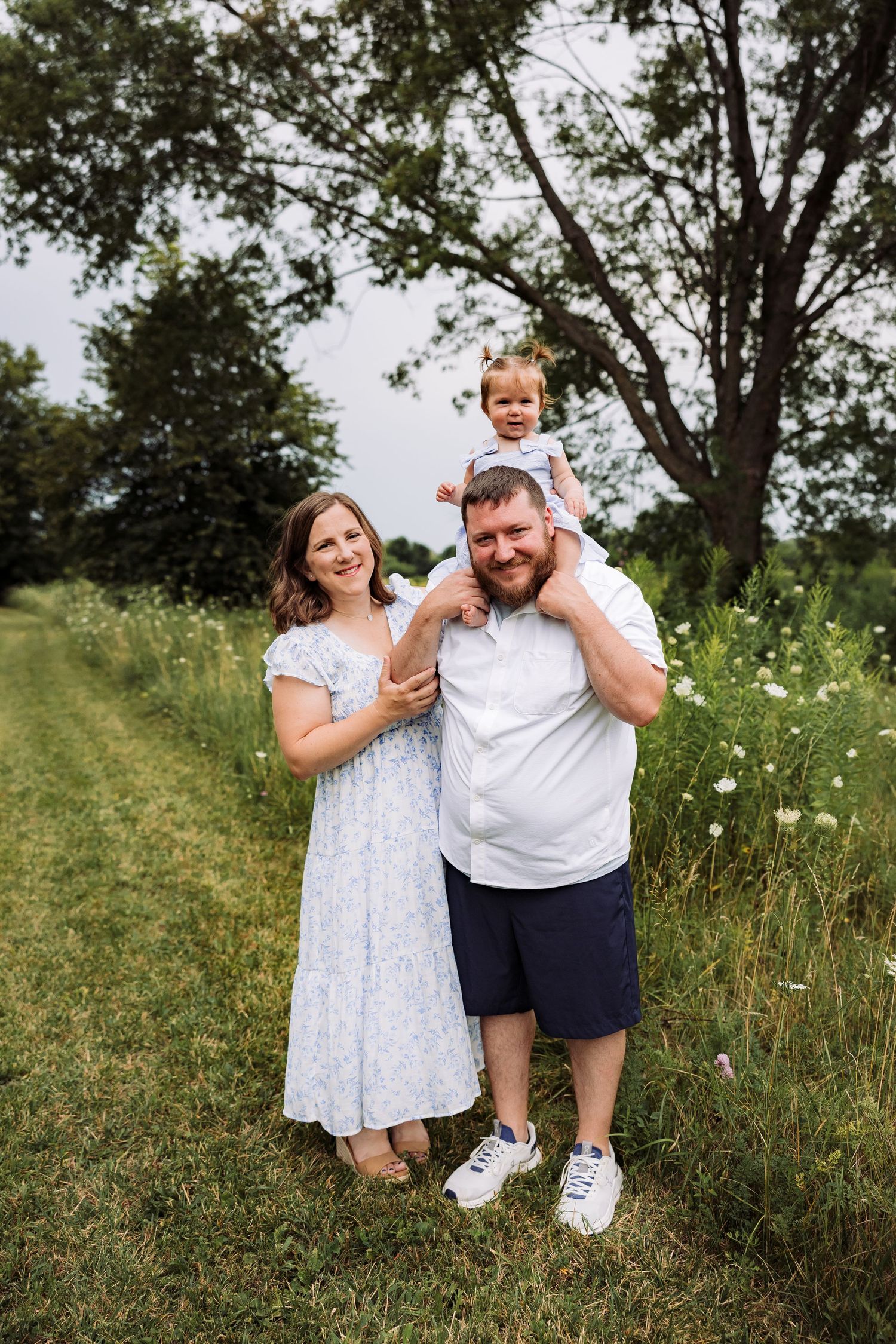A happy family of three poses outdoors, with a toddler sitting on dad's shoulders while mom stands beside them smiling.