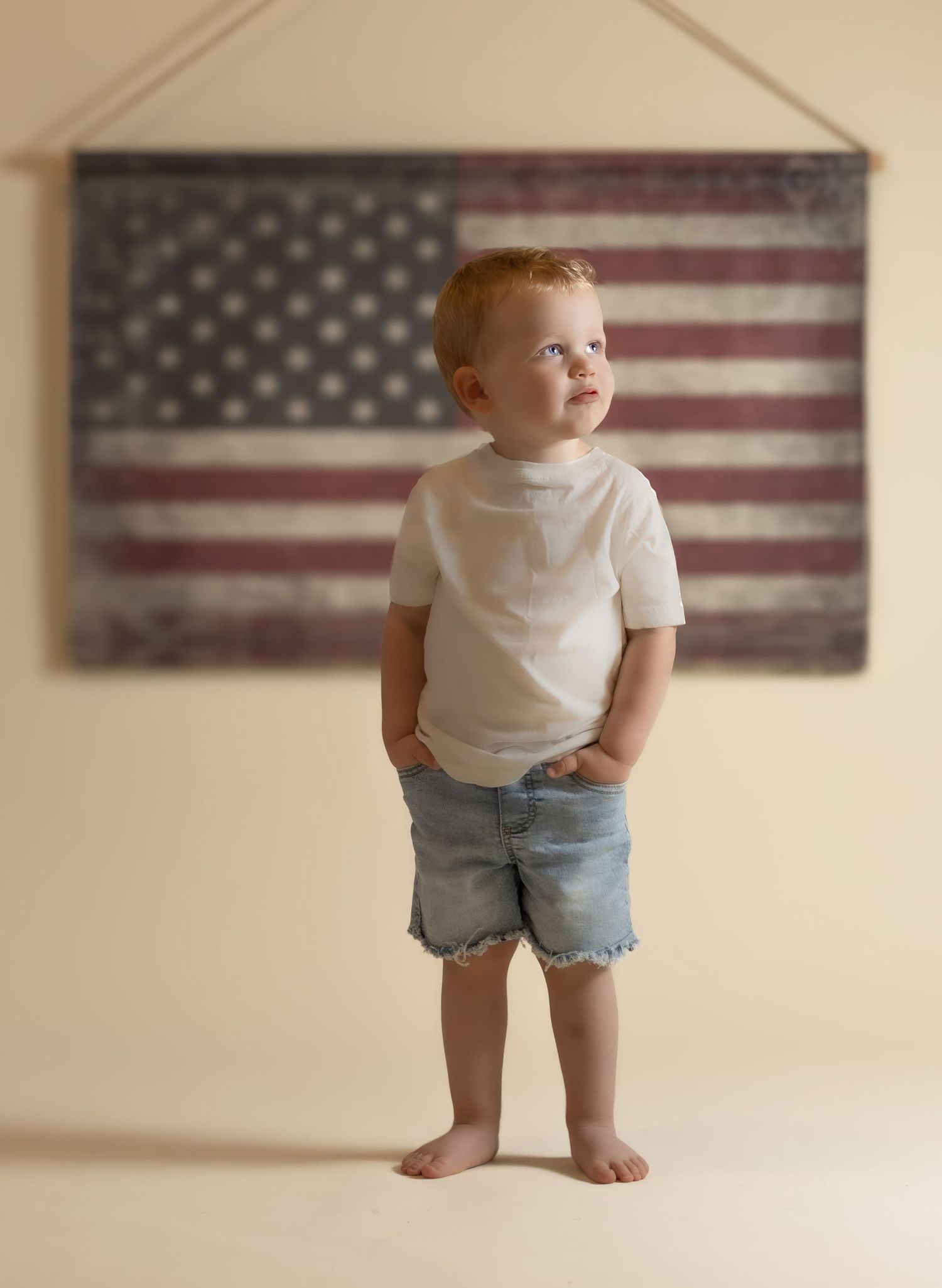 Toddler boy in white tee and denim shorts stands before vintage American flag wall hanging.