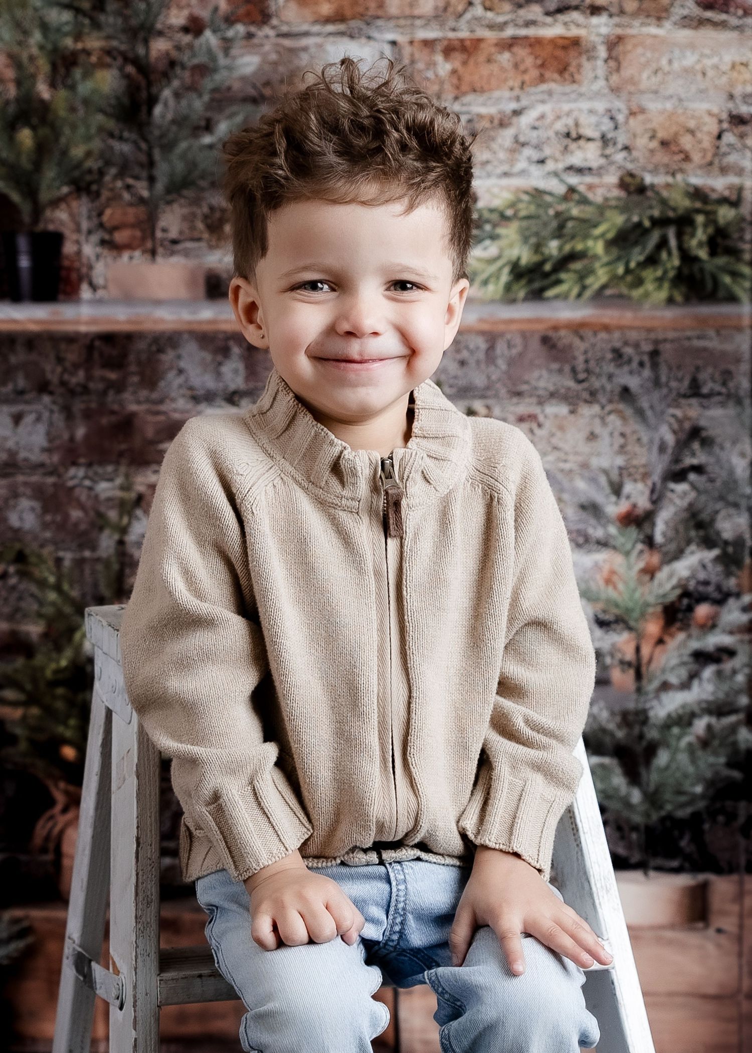 A happy young boy in a beige zip-up sweater sits smiling in front of a rustic brick wall with greenery.
