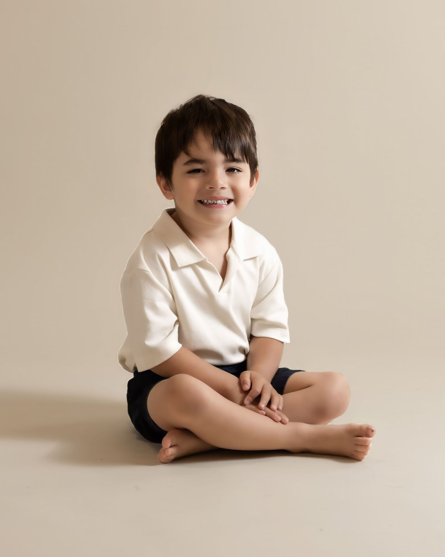 Dark-haired boy in cream polo and black shorts sits cross-legged smiling on a neutral background.