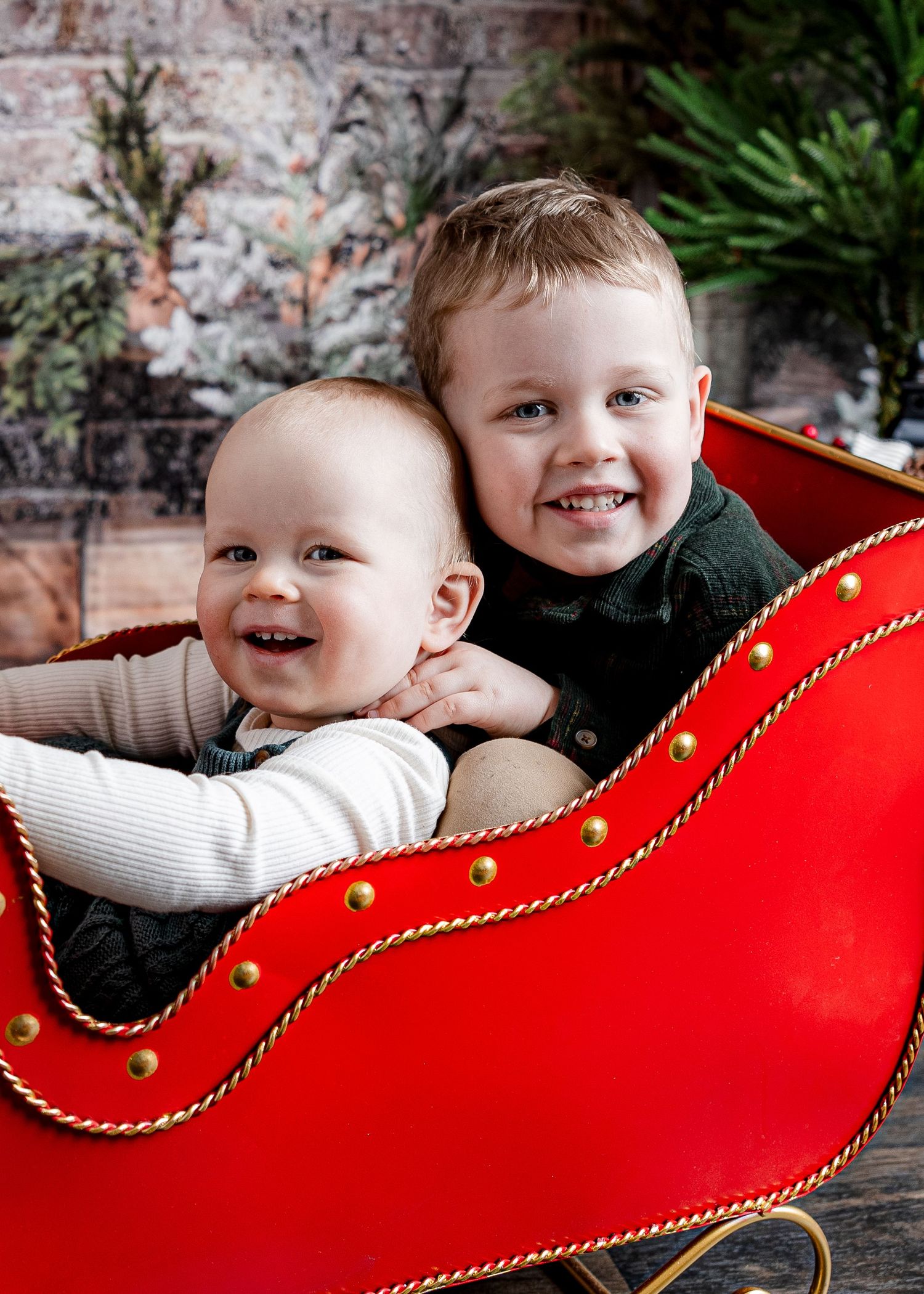 Two young brothers smiling together in a festive red Christmas sleigh with holiday decor backdrop.