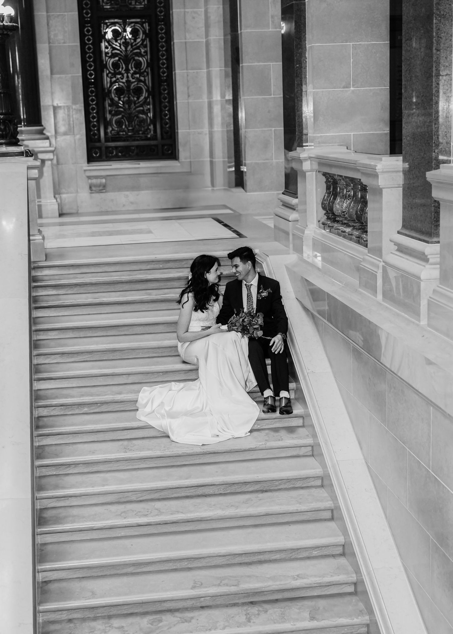 Black and white photo of bride and groom sitting closely together on grand marble staircase steps.