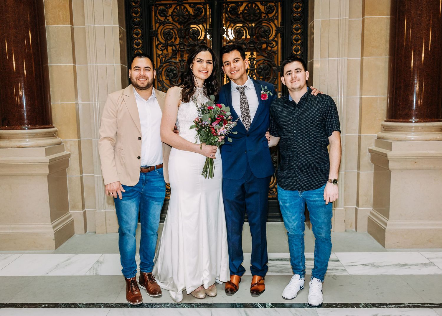 A bride holding a bouquet and groom in blue suit pose with two men before ornate golden doors.