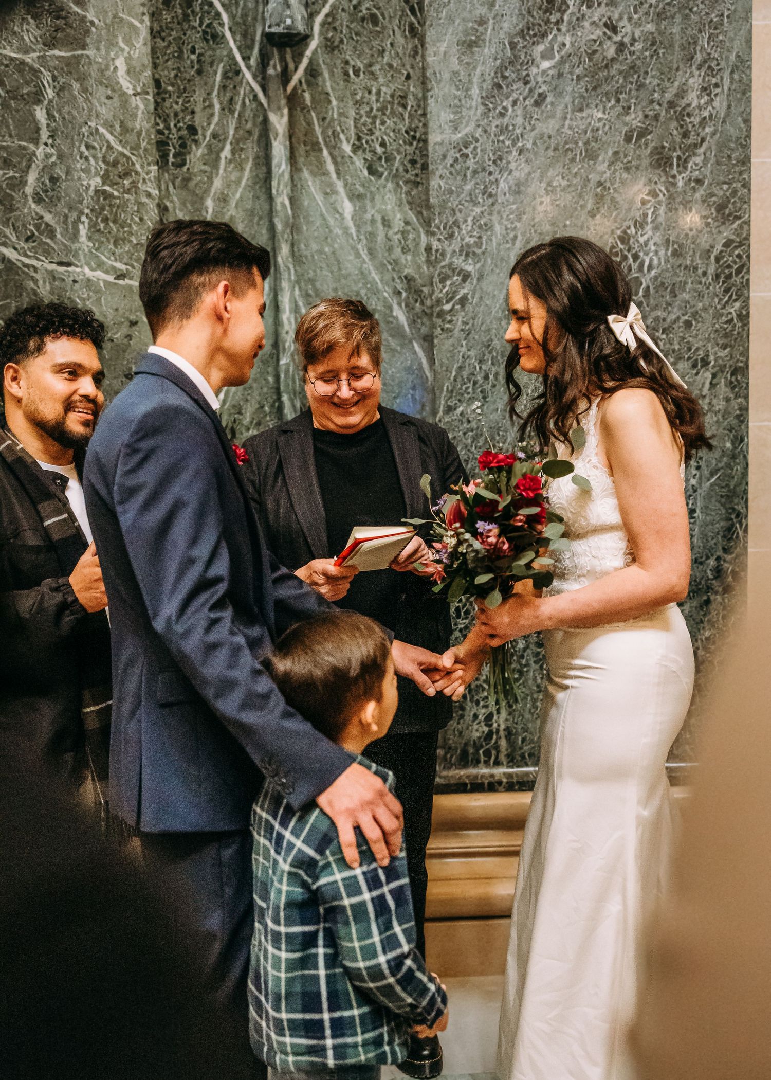 Bride and groom sharing joyful moment with officiant and young child at marble ceremony venue.