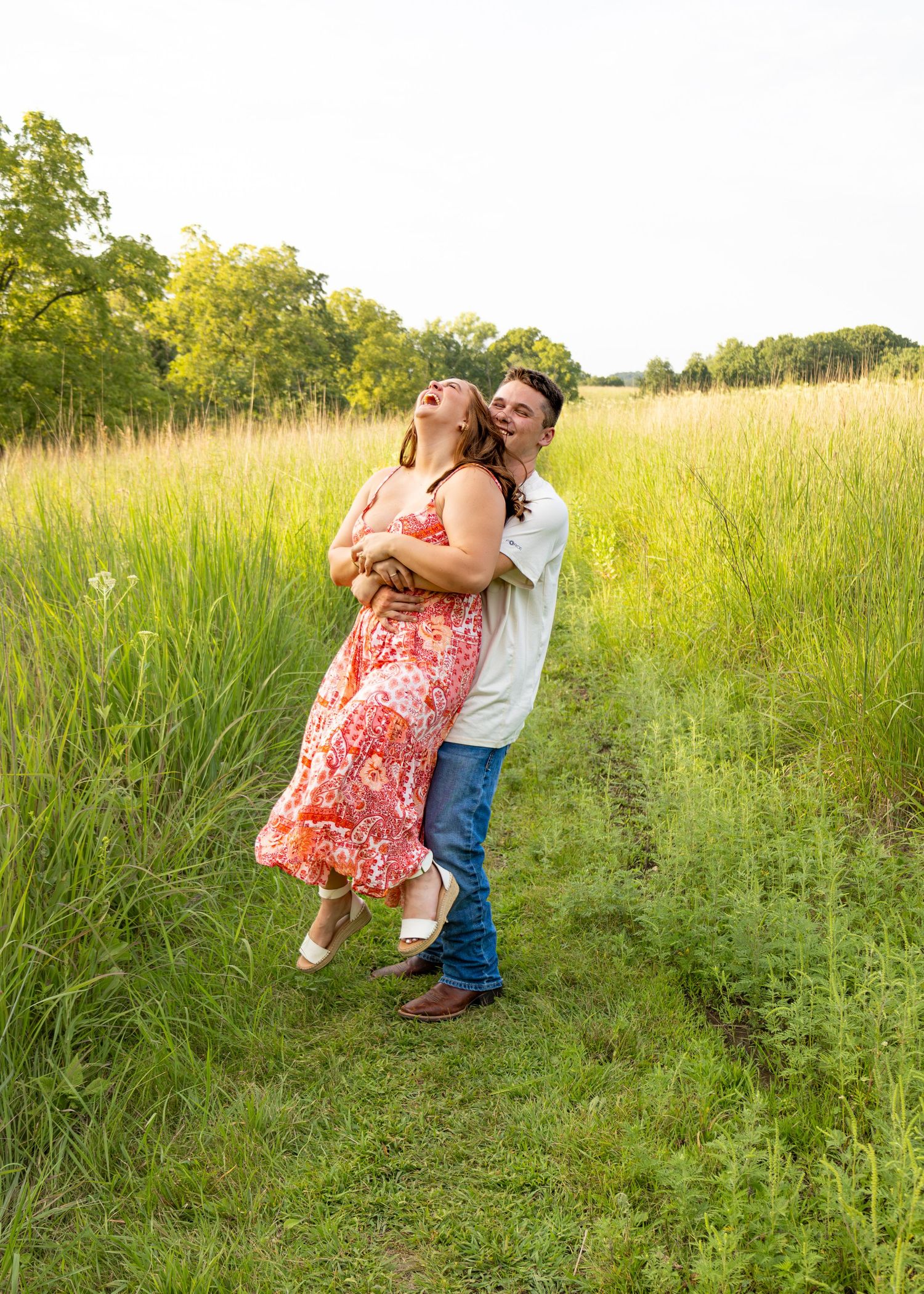 Playful couple embracing on a meadow trail at golden hour, woman in red floral dress laughing joyfully.