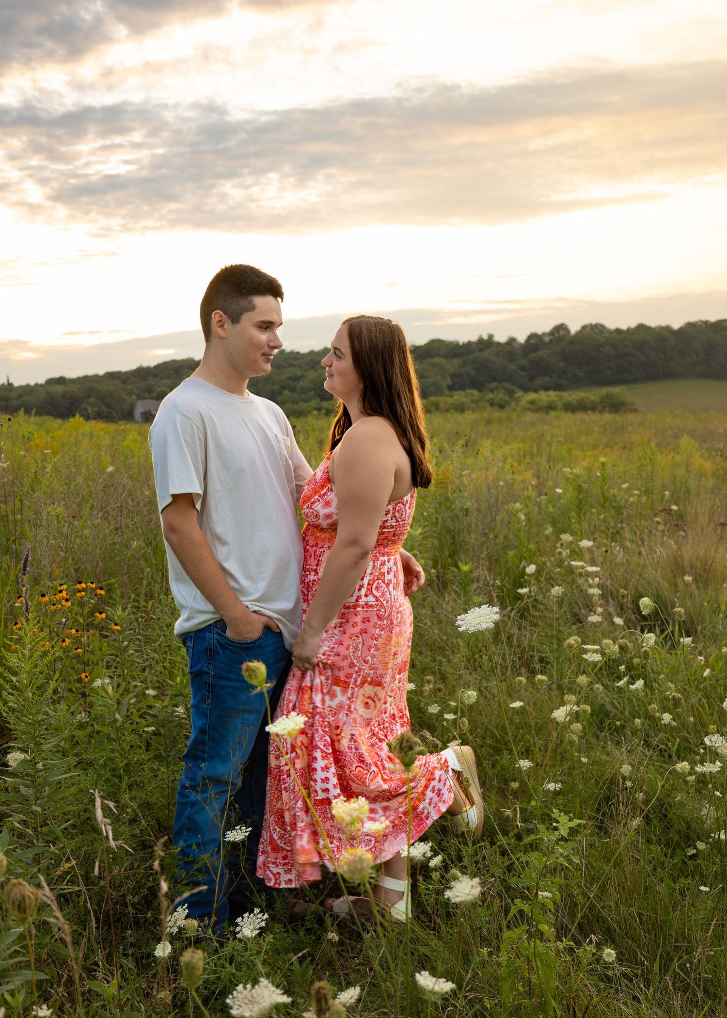 Romantic couple standing together in a wildflower field at golden hour sunset, smiling at each other lovingly.