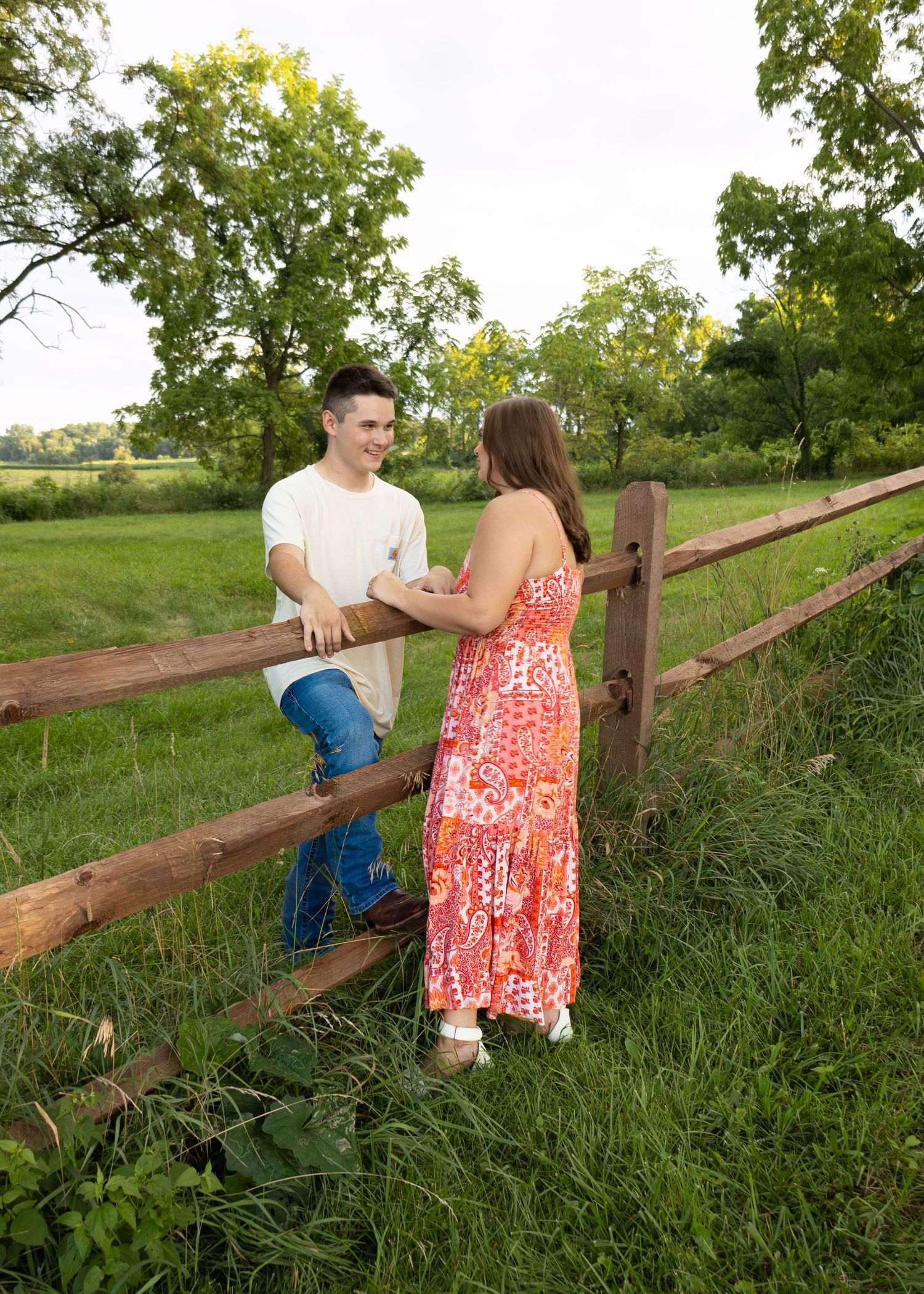 Smiling young couple standing by rustic wooden fence in green pastoral landscape on cloudy day.
