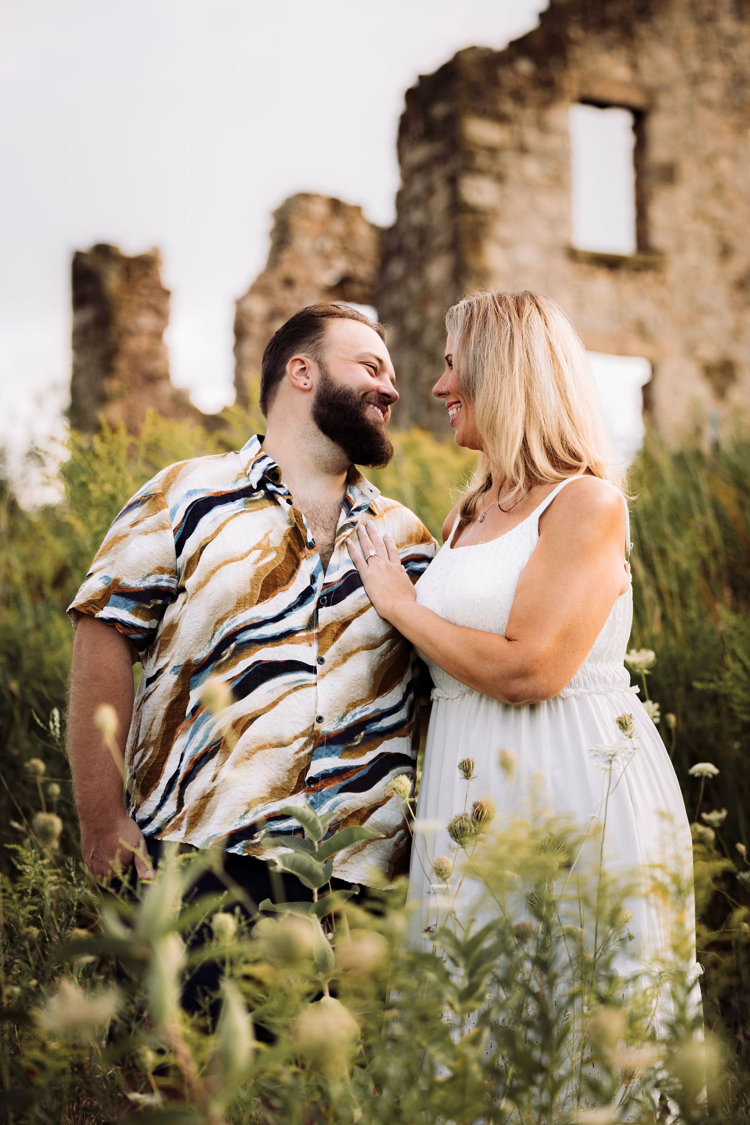 A smiling couple stands among wildflowers near stone ruins, woman in white dress, man in colorful shirt.