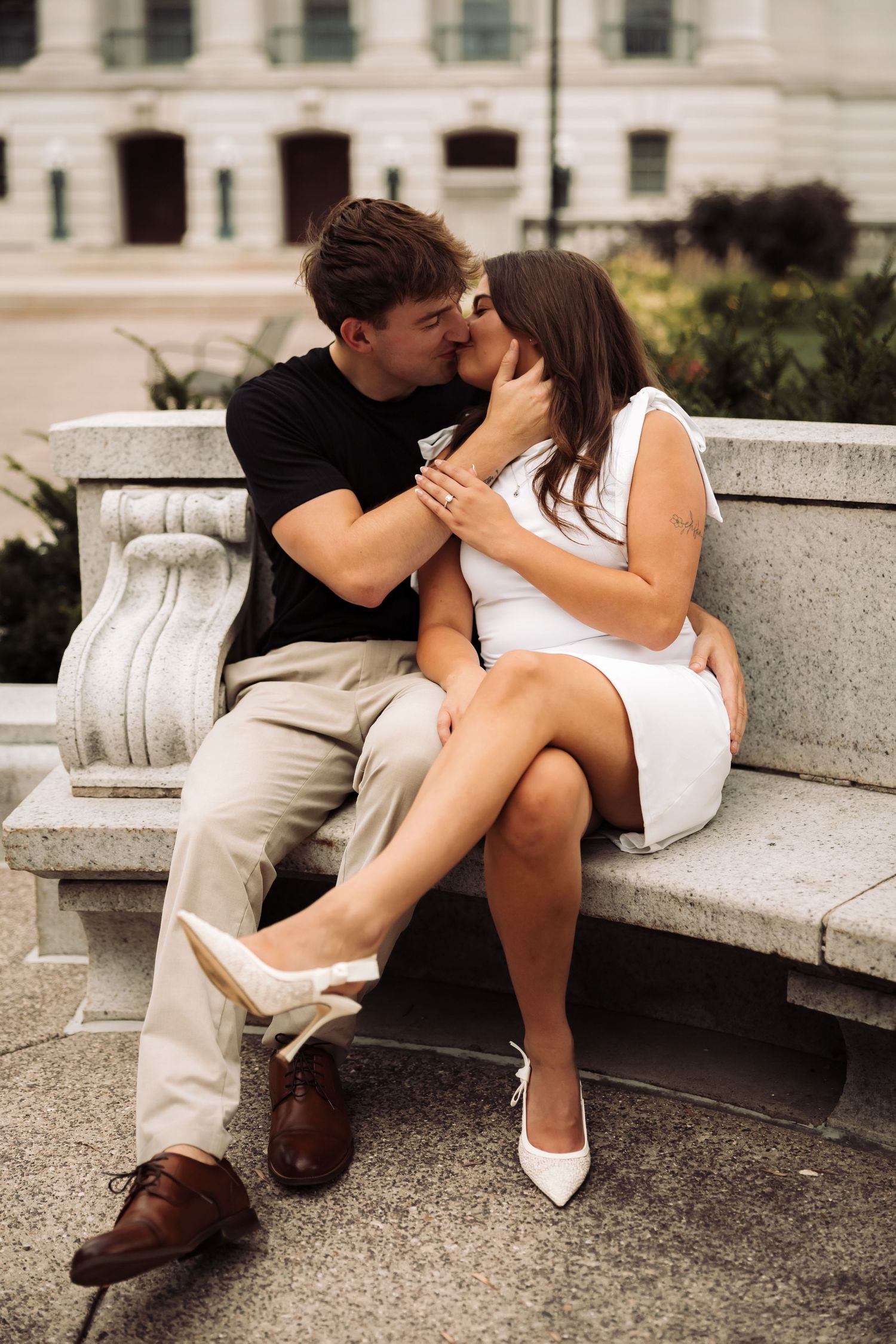 Romantic couple sharing a kiss on a stone bench outdoors, woman in white dress and heels, man in black shirt and khakis.