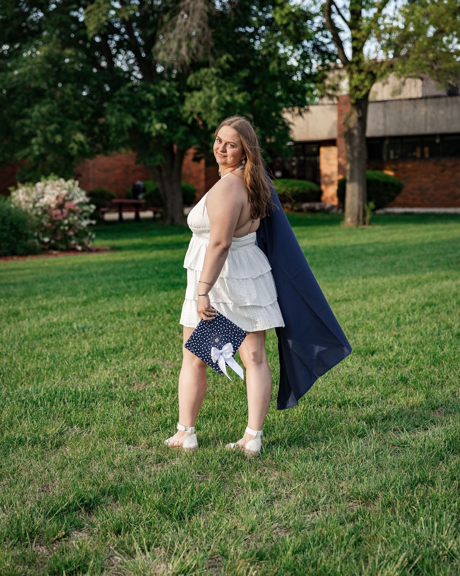 Young woman in white ruffled top and polka dot shorts holding navy blazer, standing on green lawn near brick building.