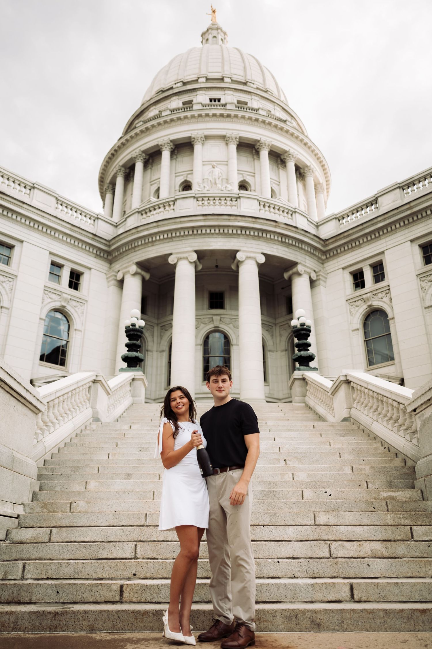 Couple posing on steps of white neoclassical capitol building with dome and columns on cloudy day.