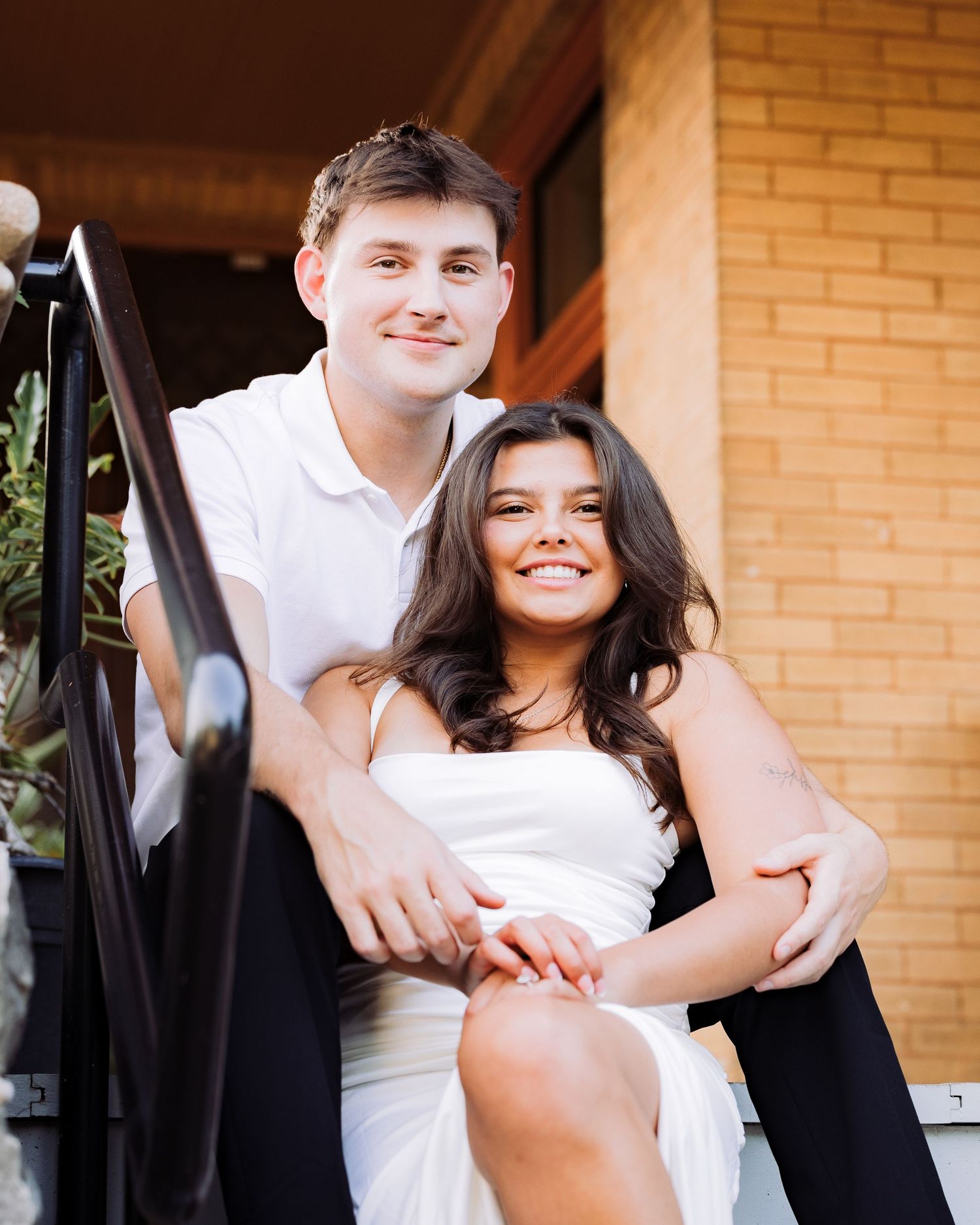 A smiling young couple in white outfits sitting together on outdoor steps in front of a brick building.