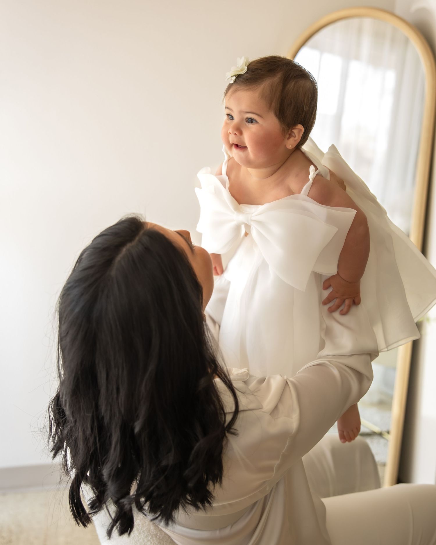 A dark-haired mother in white lifts her smiling baby girl in a white bow dress near a gold mirror.