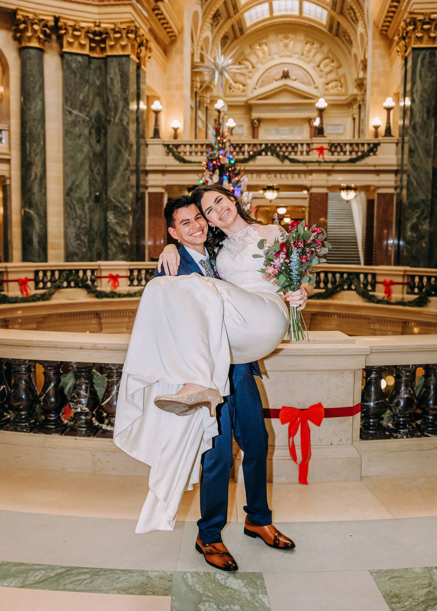 Groom carries bride in colorful wedding photo inside grand capitol rotunda with holiday decorations.