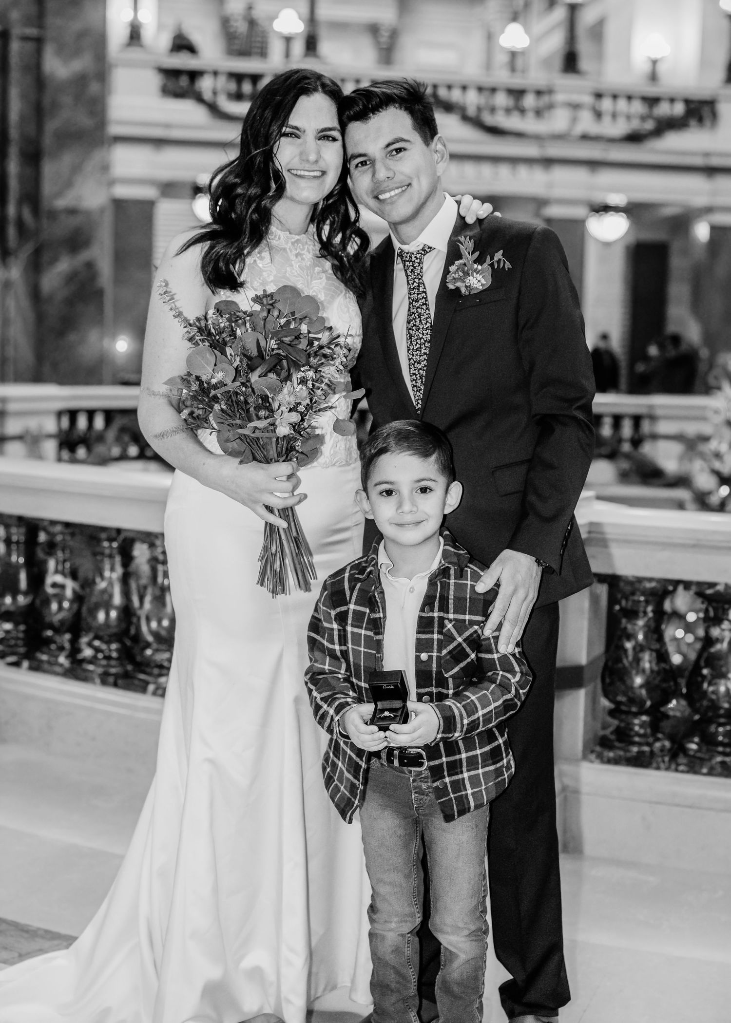Wedding couple in formal attire poses with young boy holding ring box in grand marble venue.
