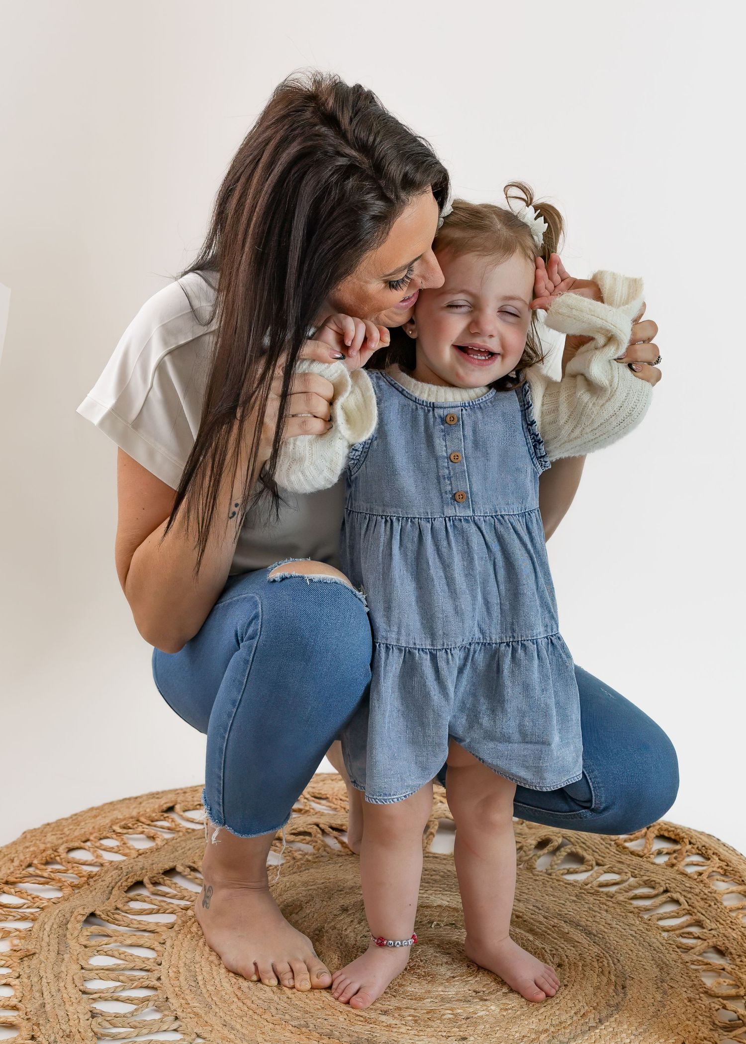 Mother kissing laughing toddler daughter in denim dress during indoor photo session on woven rug.