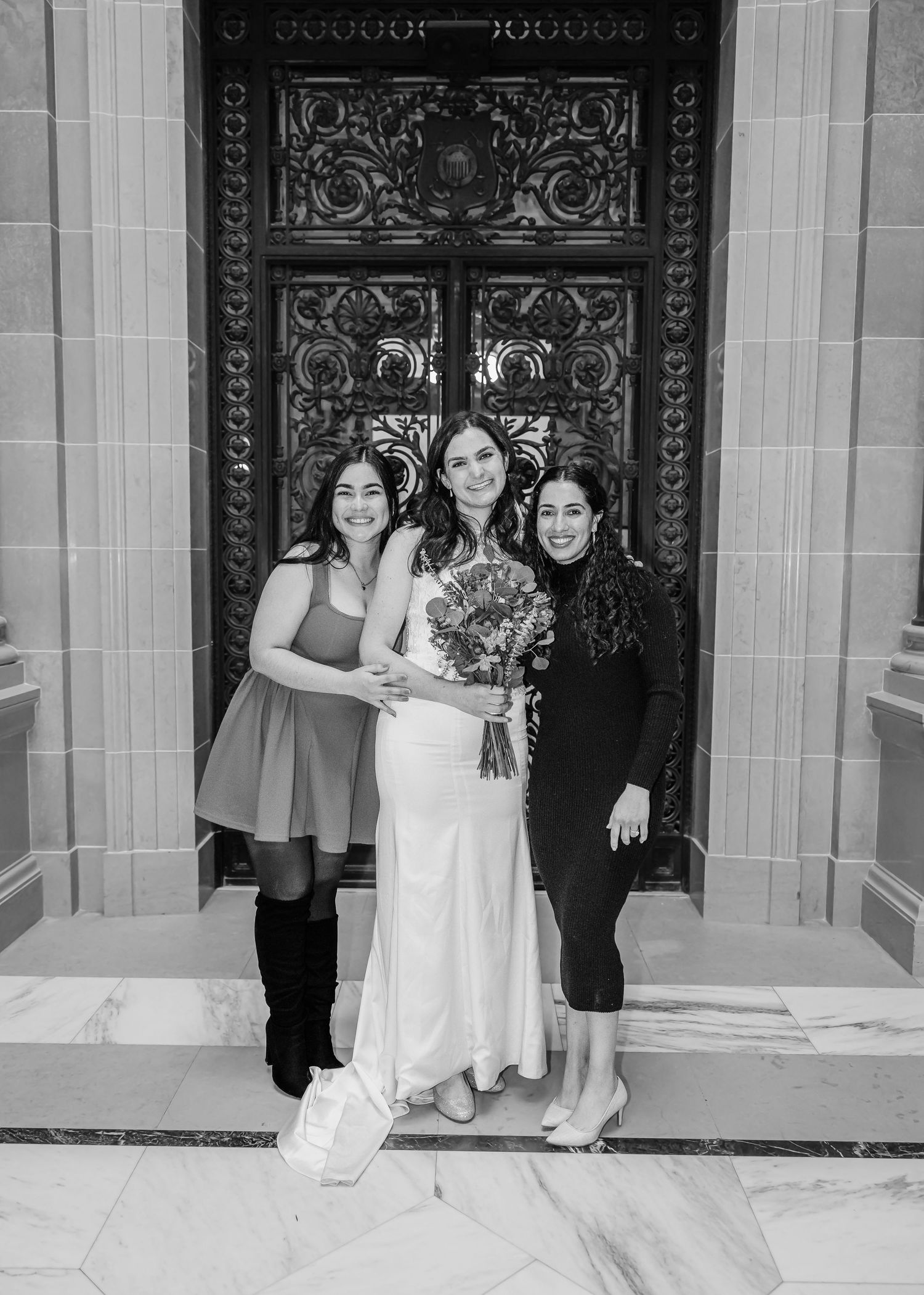 Bride holding bouquet standing between two women before ornate doors in black and white photo.