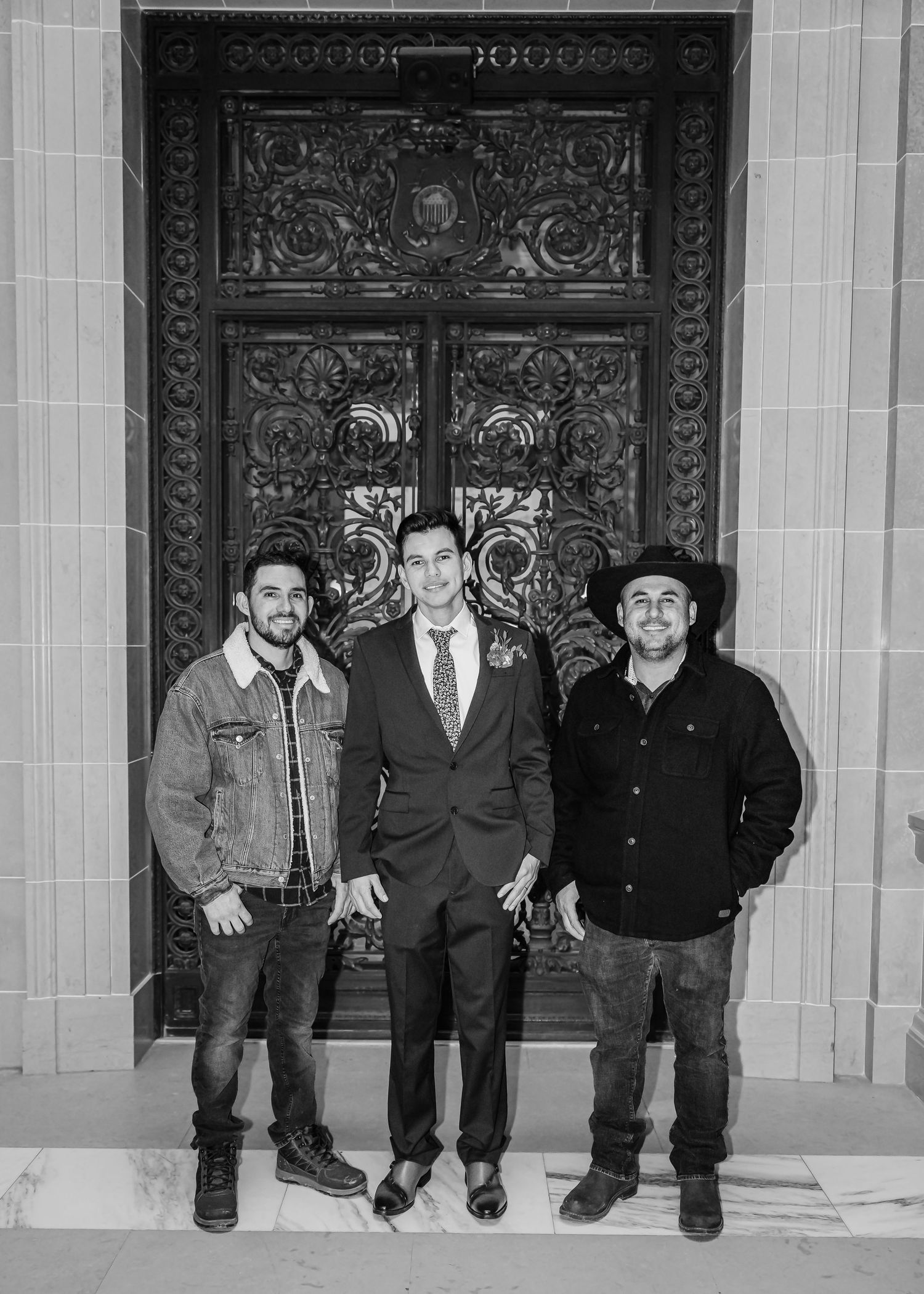 Black and white portrait of groom flanked by two male guests before an ornate carved wooden door.