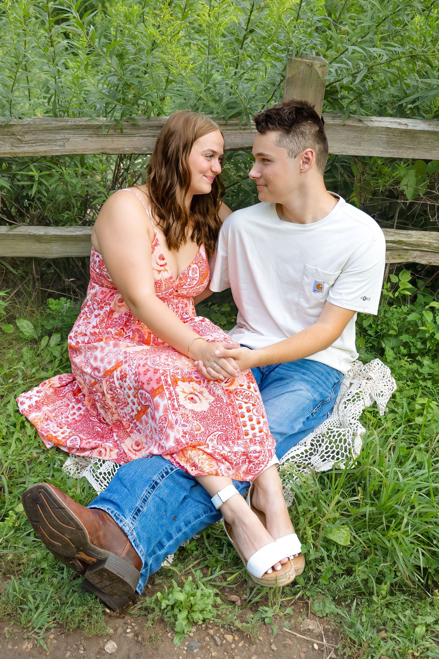 A young couple sits together on a lace blanket by a wooden fence, gazing into each other's eyes outdoors.