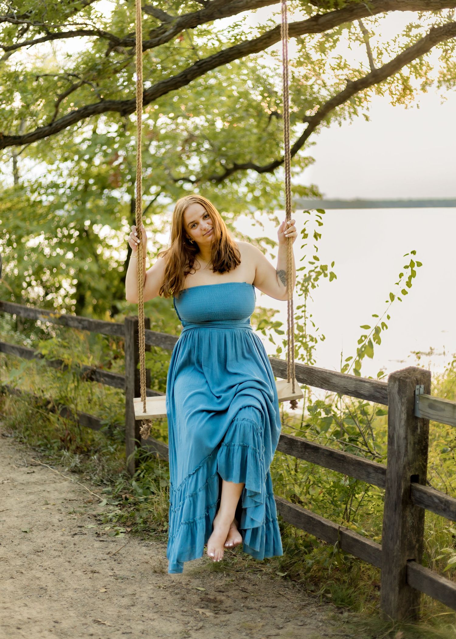 Young woman in blue strapless maxi dress sitting on wooden rope swing near lakeside fence at sunset.