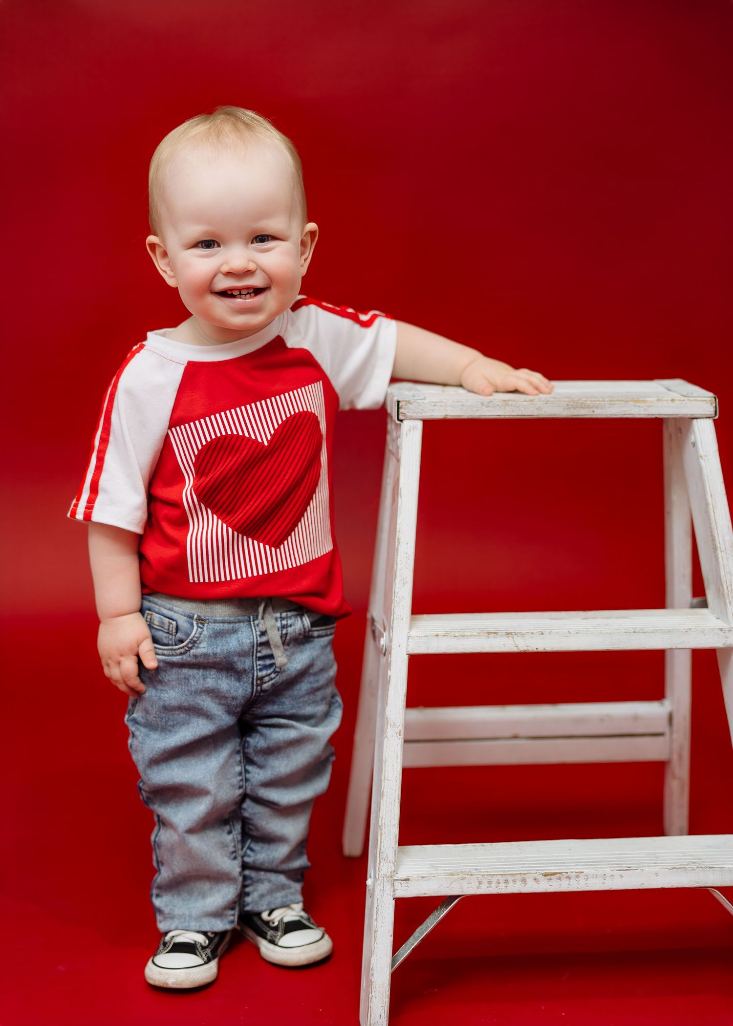 Smiling toddler in red heart shirt stands beside white ladder on red background.