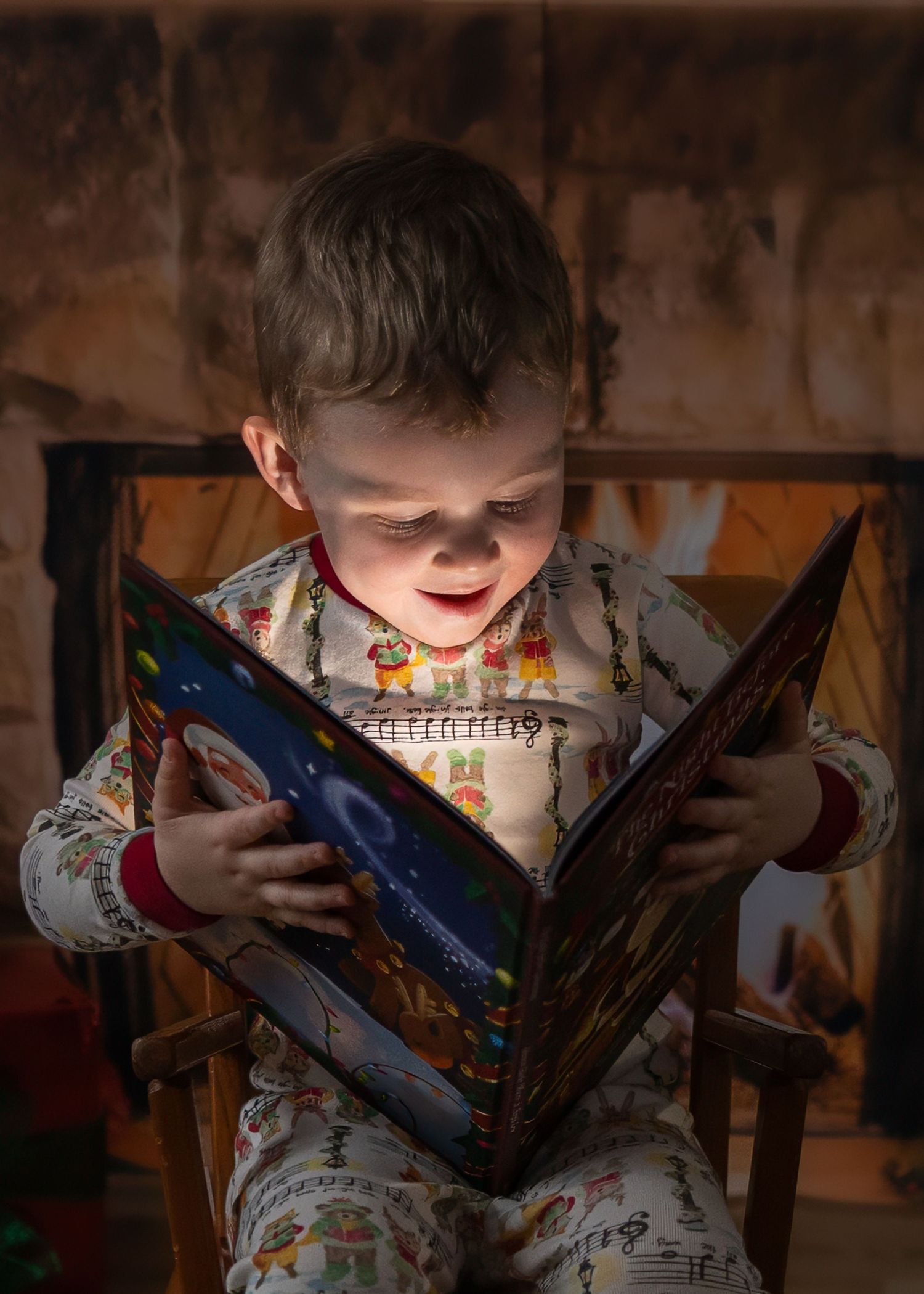 Young child in holiday pajamas reading a glowing Christmas book in a dimly lit room.