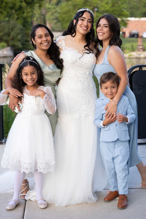 Smiling bride in a white lace gown and tiara poses outdoors with two bridesmaids, a flower girl in a white dress, and a ring bearer in a lig
