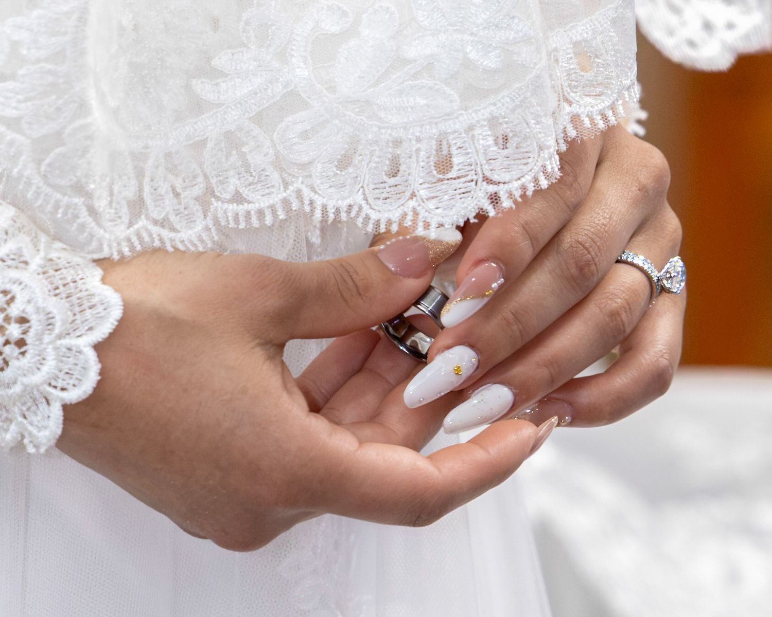 Bride in a white lace gown clasping her hands, showing a diamond engagement ring and long white manicured nails with gold accents.