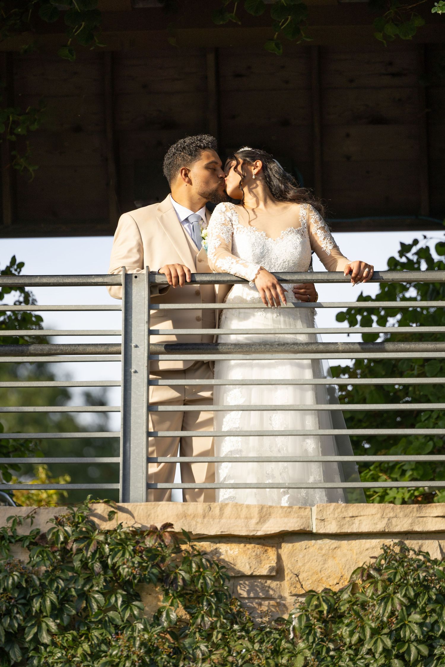 A bride in a lace wedding gown and a groom in a tan suit share a kiss on a balcony with a metal railing, surrounded by green ivy.