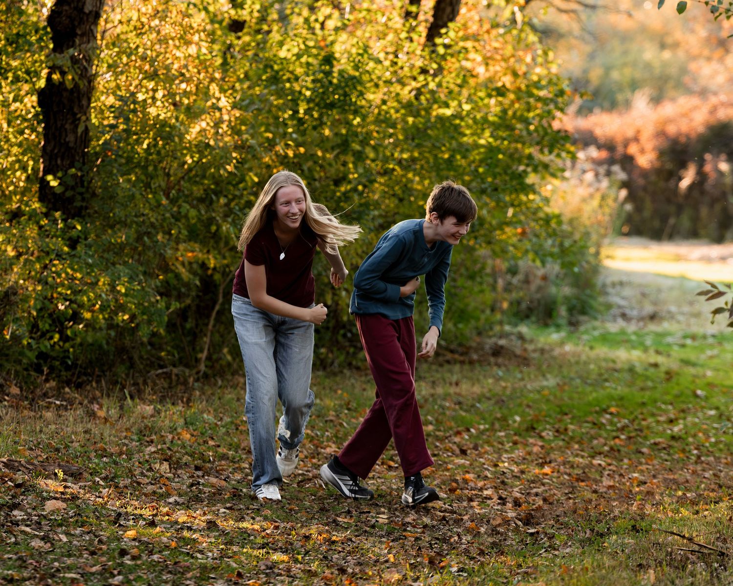 Two kids laugh and play together on a grassy path in an autumn park, surrounded by golden fall foliage and sunlit trees.