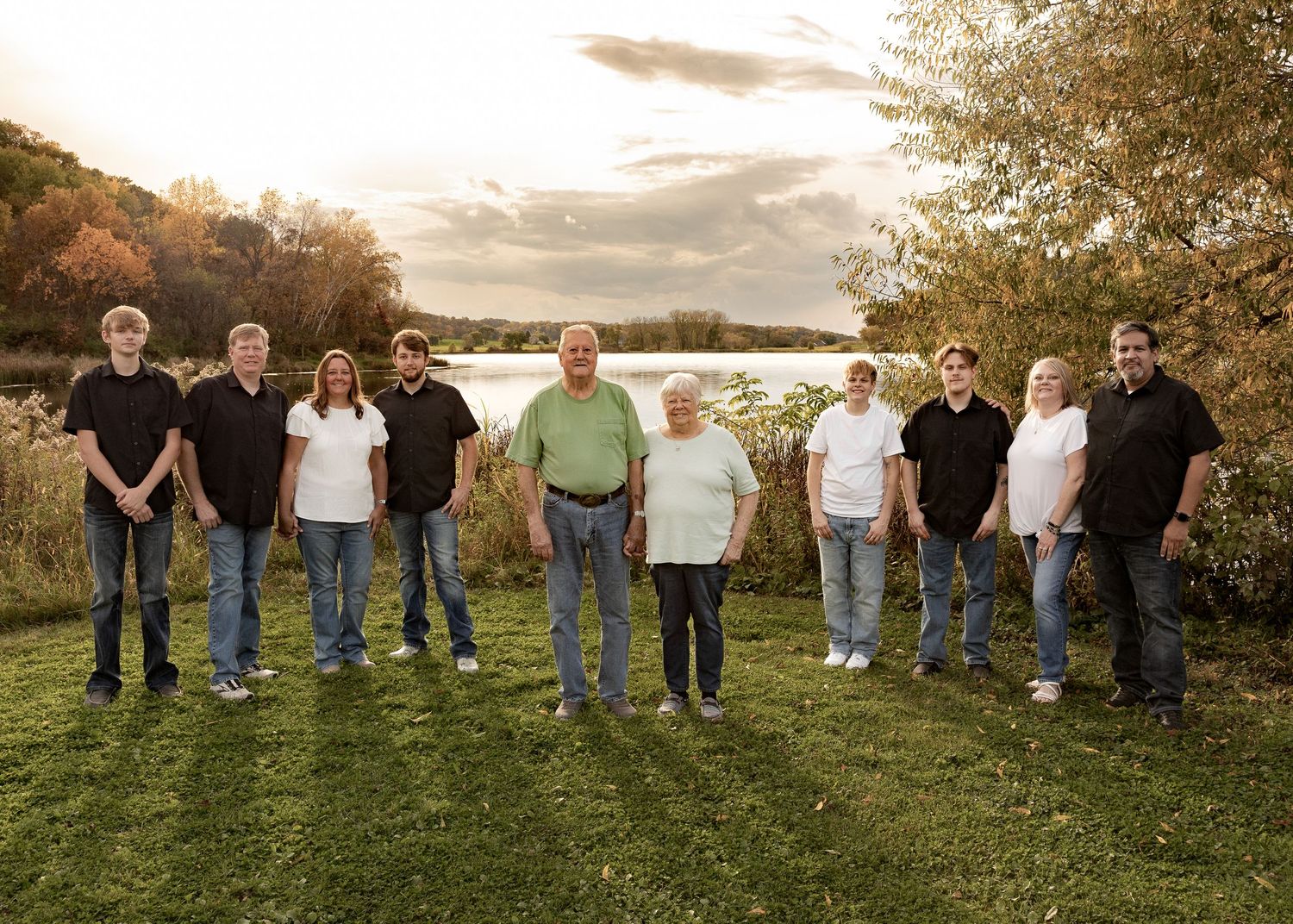 Large family group standing outdoors near scenic lake with autumn trees.
