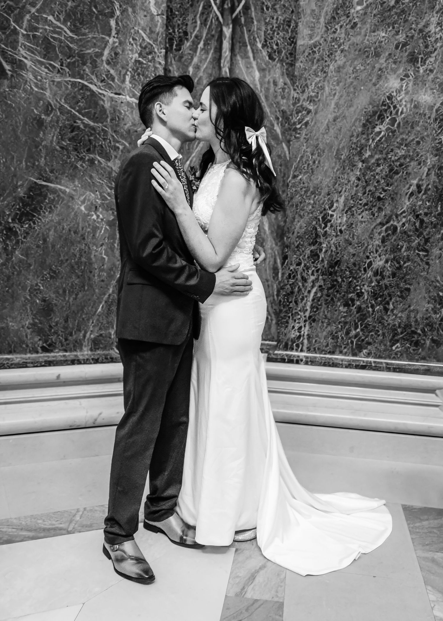 Bride and groom share first kiss in black and white wedding portrait before marble backdrop.