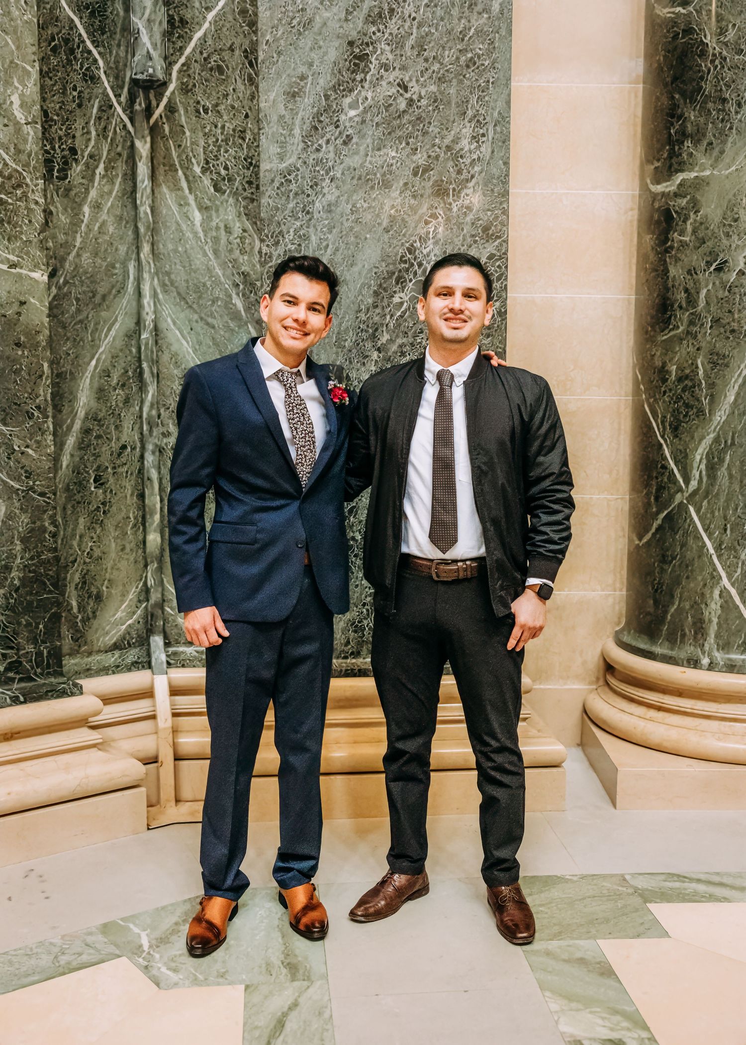 Two smiling men in suits standing together in front of marble columns at a wedding venue.