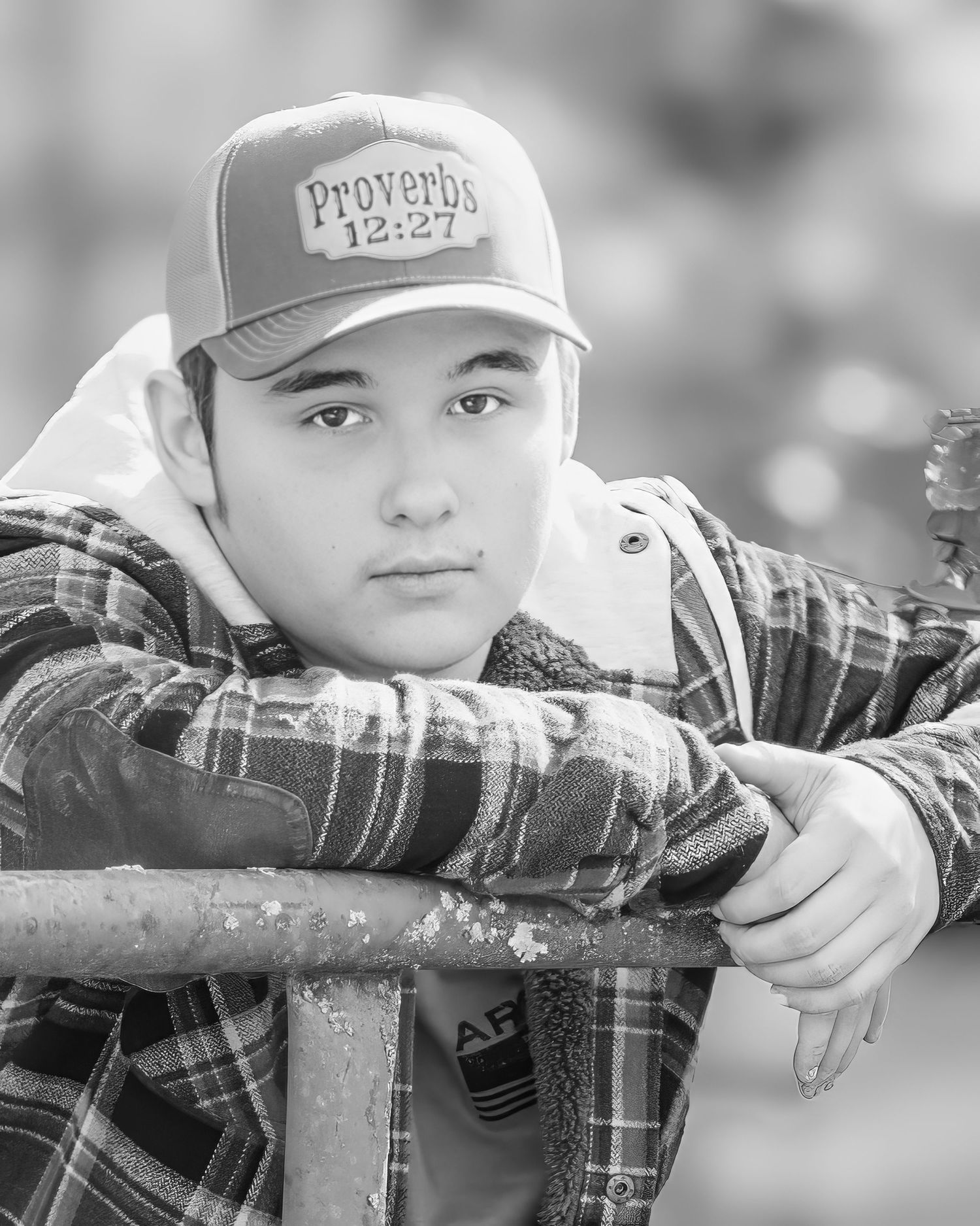 Black and white close-up senior portrait of teen boy in Proverbs cap leaning on rustic railing.
