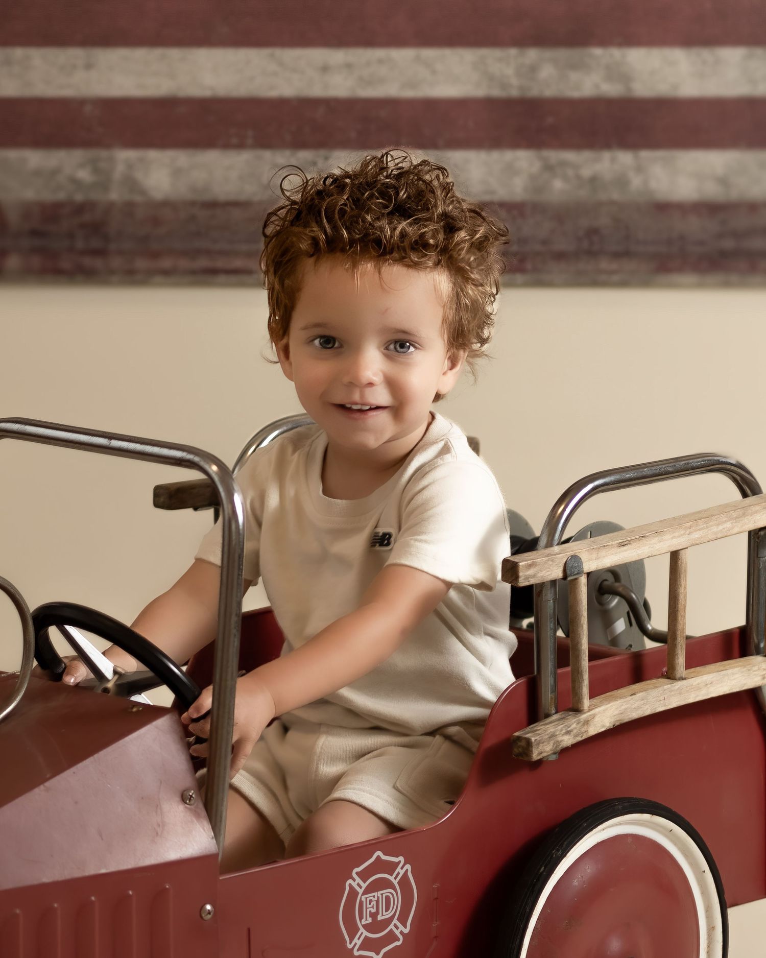 Curly-haired toddler smiling in a vintage red fire truck pedal car indoors.