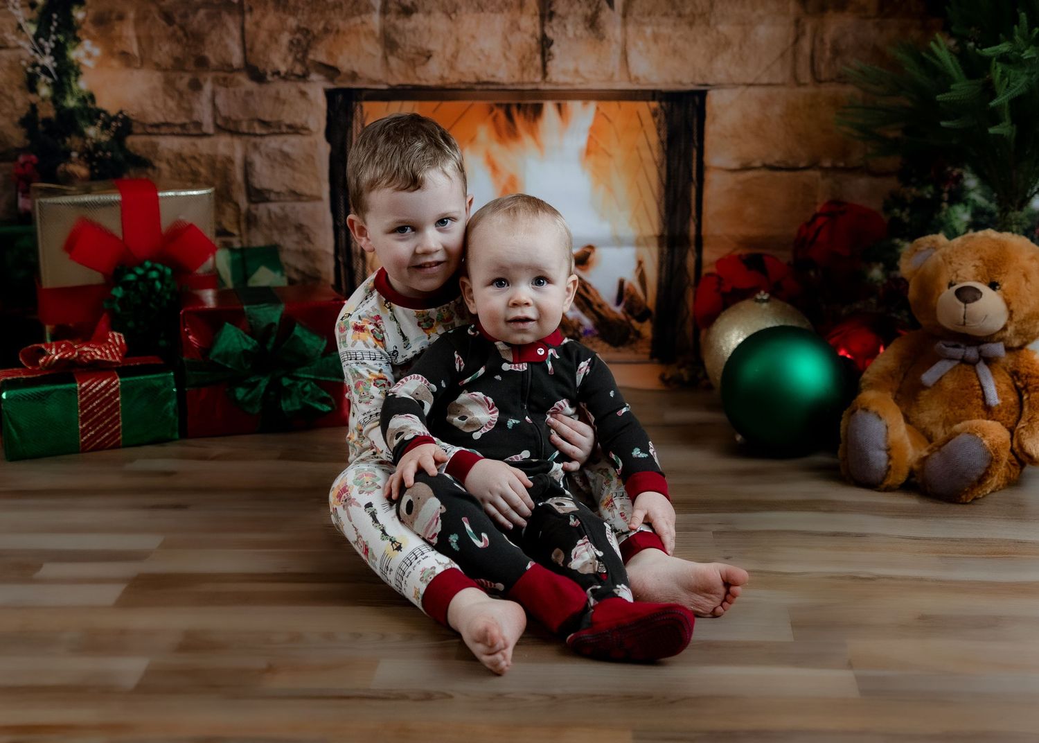 Two young brothers in Christmas pajamas sitting together by a decorated fireplace with holiday gifts.