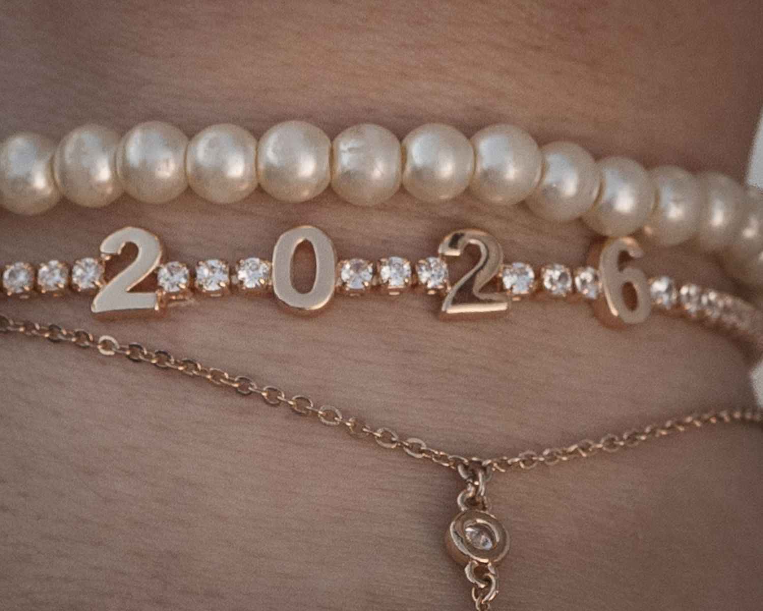 Three stacked bracelets featuring pearl, rhinestone with gold 2026 numbers, and delicate gold chain designs.