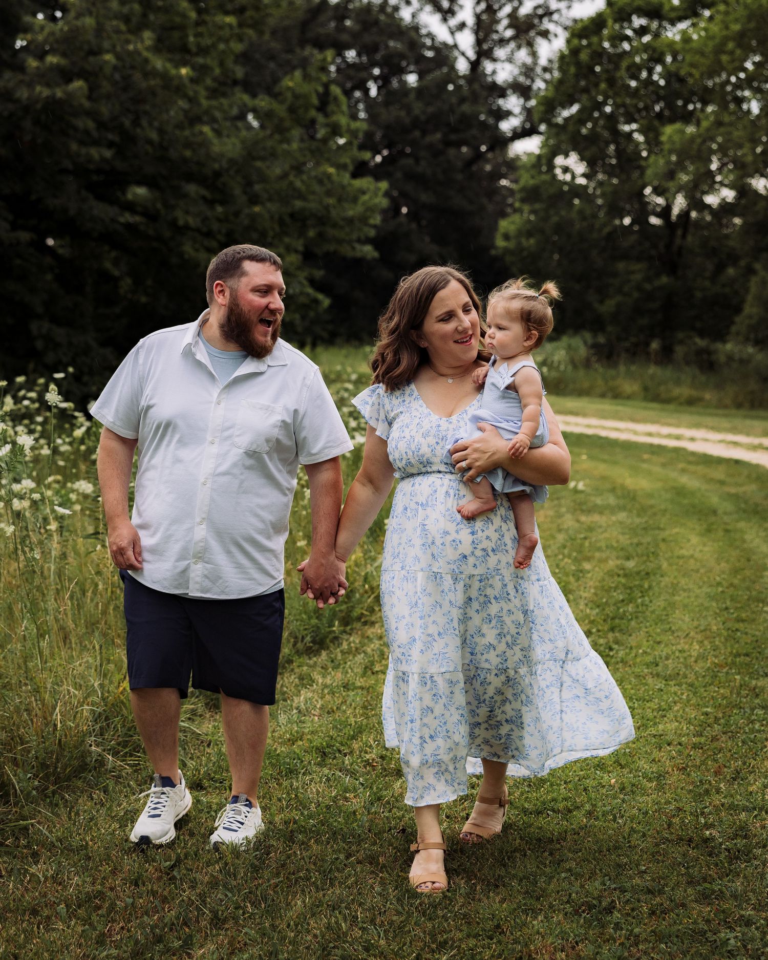 A happy family of three walks hand-in-hand through a lush green meadow, with a woman holding a baby in a blue floral dress.