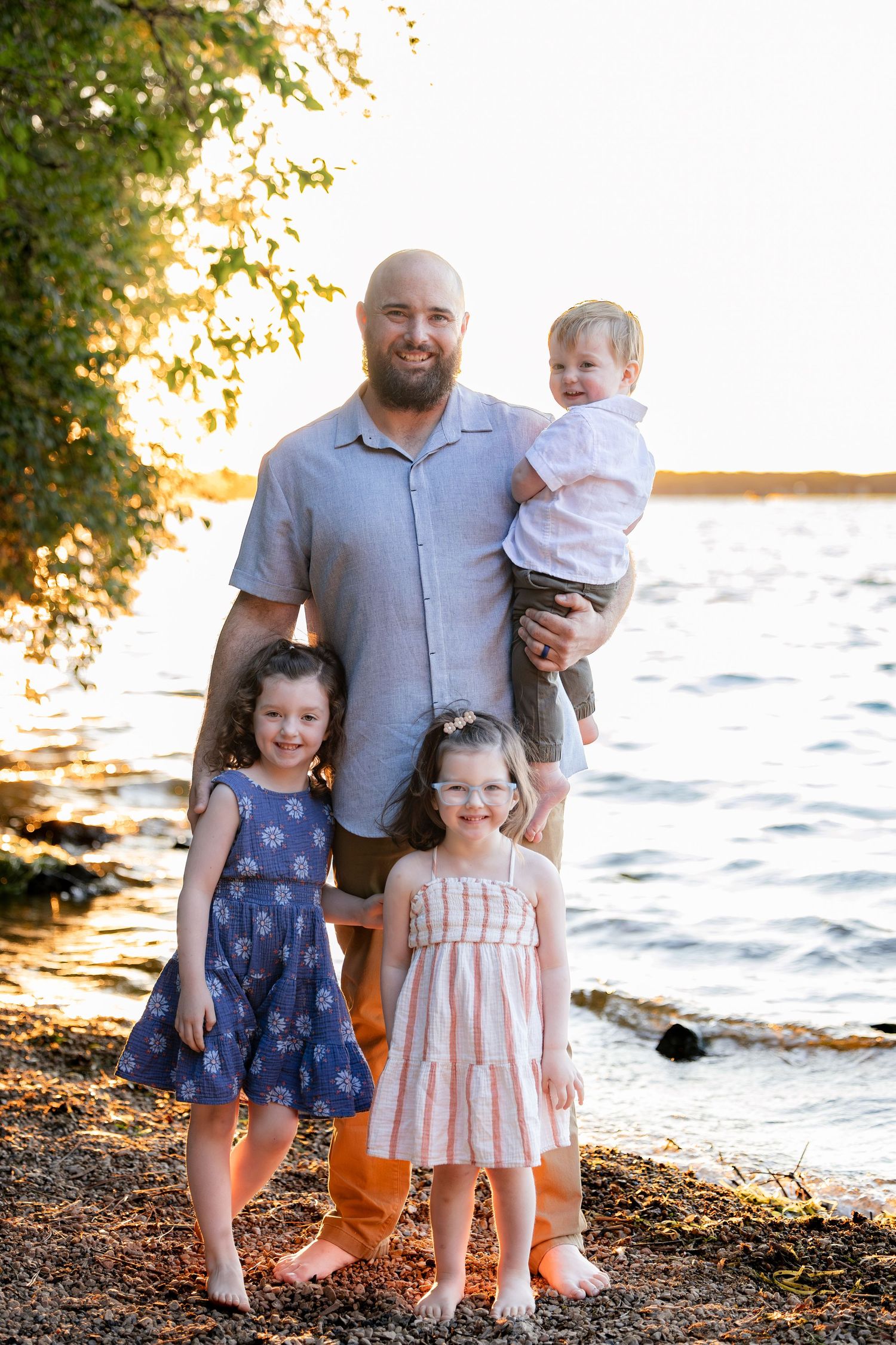 Bald bearded father holds toddler boy while two young girls stand in front, posing by a sunlit lake shore.