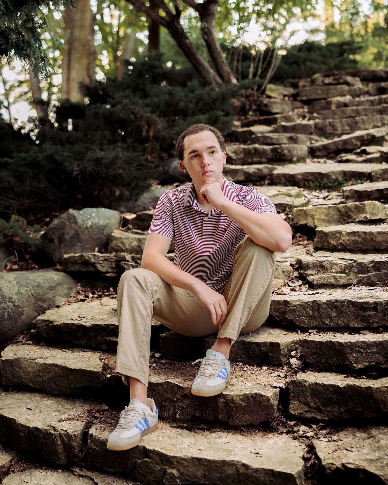 Young man in pink polo and khakis sitting thoughtfully on stone steps surrounded by lush greenery.