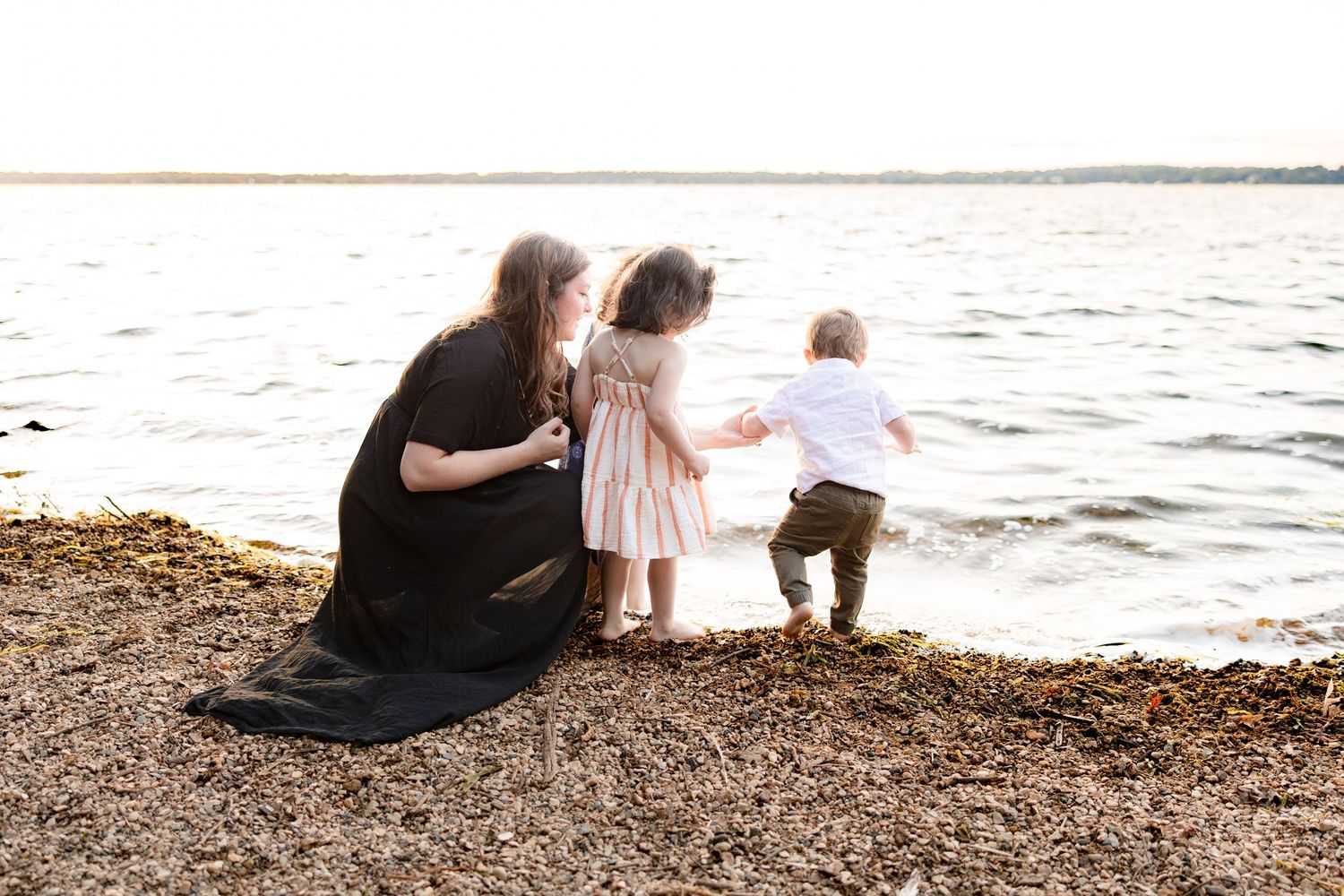 A mother in a black dress kneels with two young children by a calm waterfront at sunset.