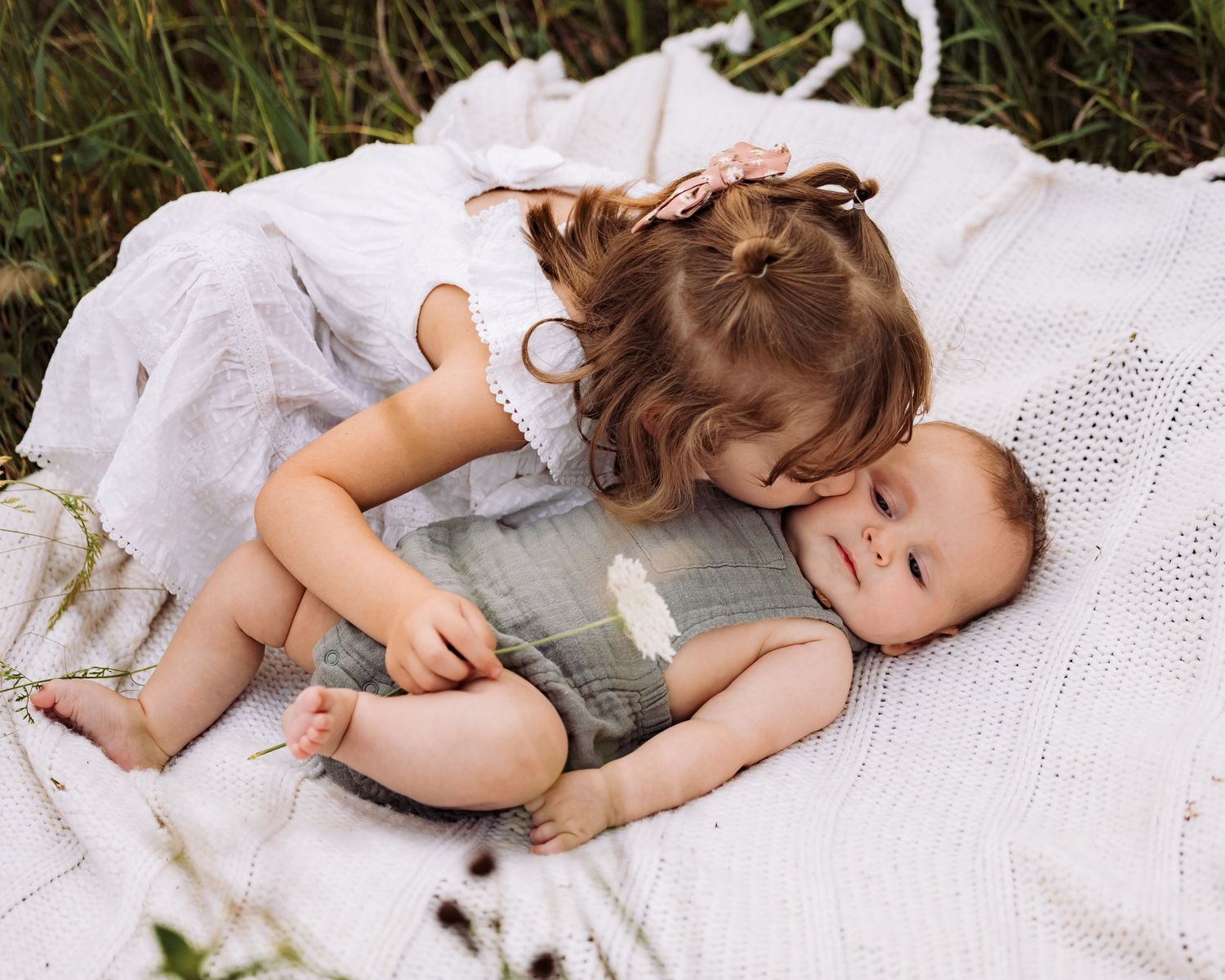 A toddler girl in a white dress cuddles her baby sibling on a white blanket in a green field.