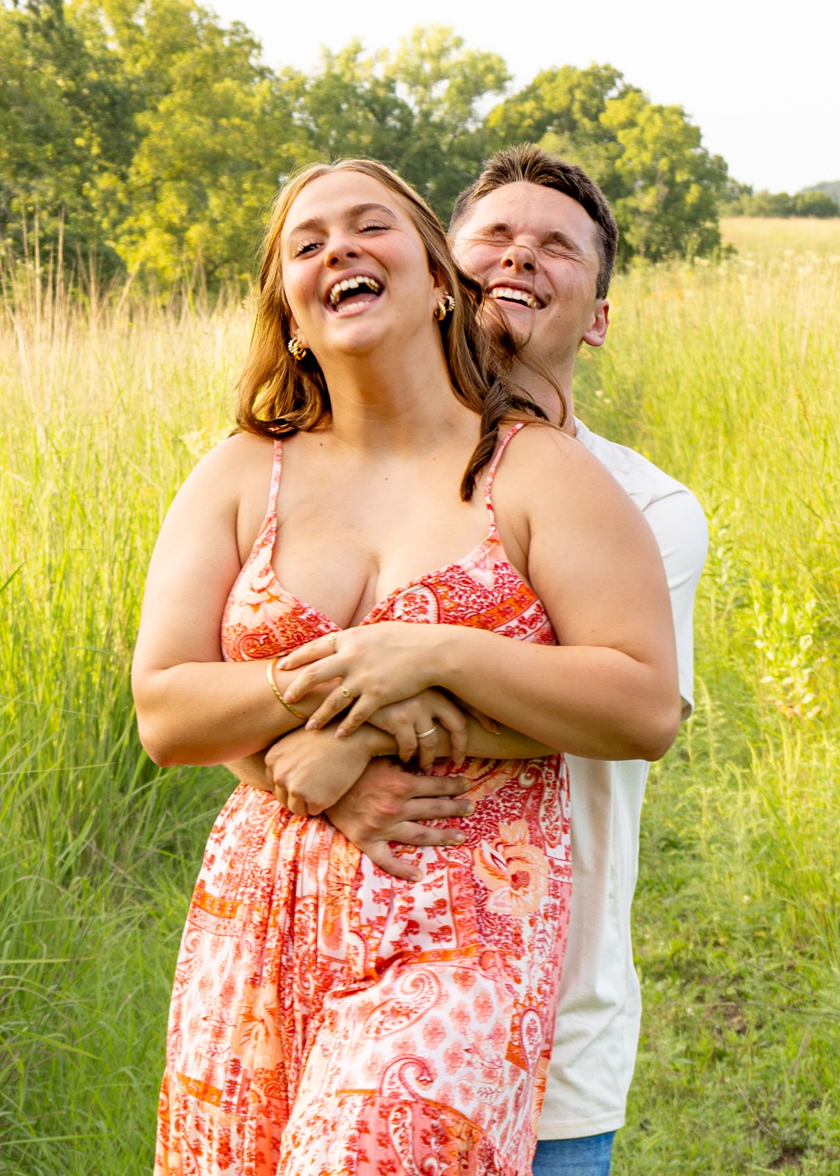 Happy couple embracing and laughing in a sunny green meadow, woman wearing a red floral sundress.