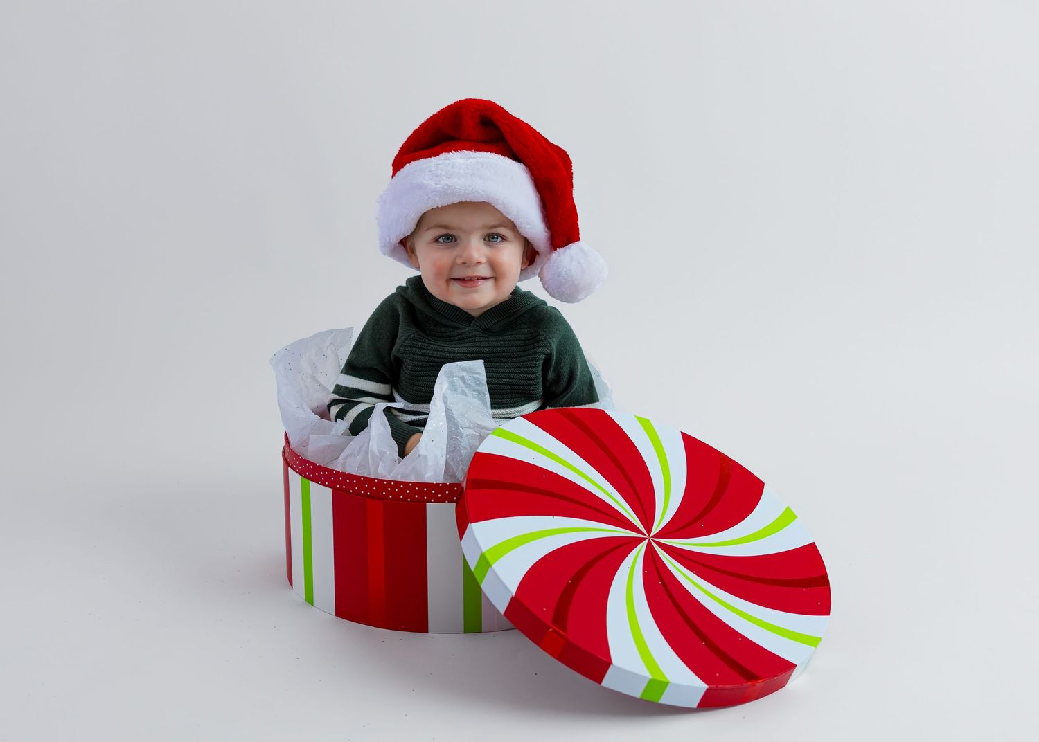 Adorable baby in Santa hat sitting inside large red and white striped Christmas gift box.