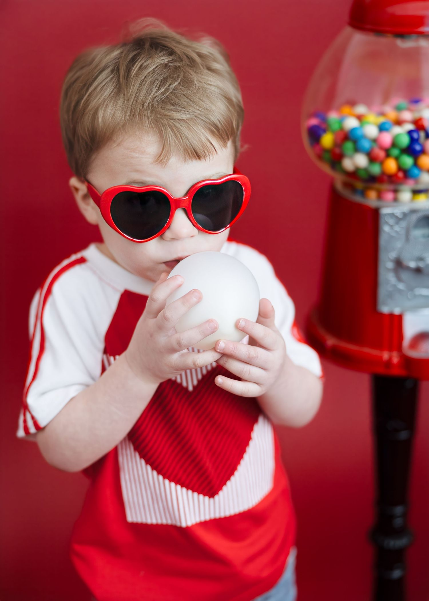 Toddler in heart sunglasses blows bubble near colorful gumball machine.