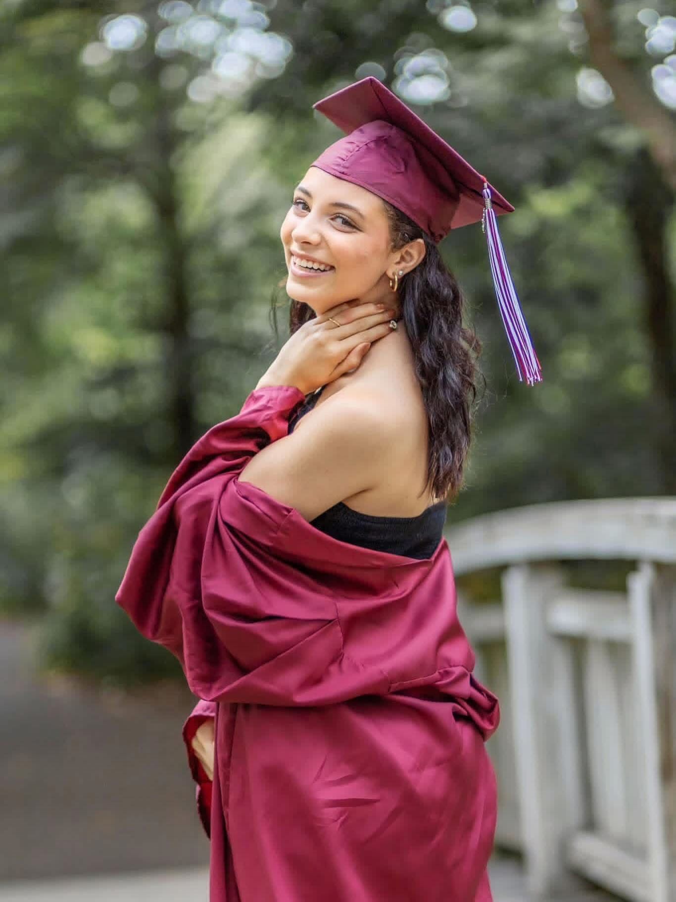 Smiling graduate in burgundy cap and gown posing outdoors among green trees for senior portrait.
