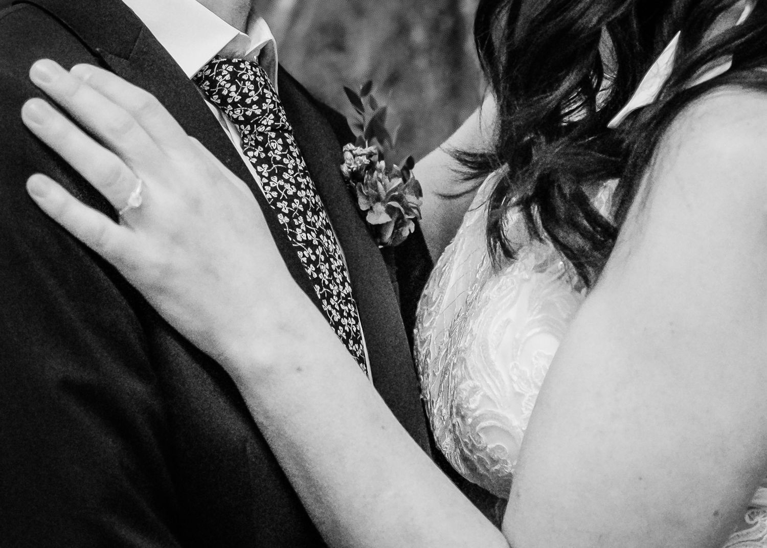 Black and white close-up of newlyweds embracing, showing wedding ring and floral boutonniere.