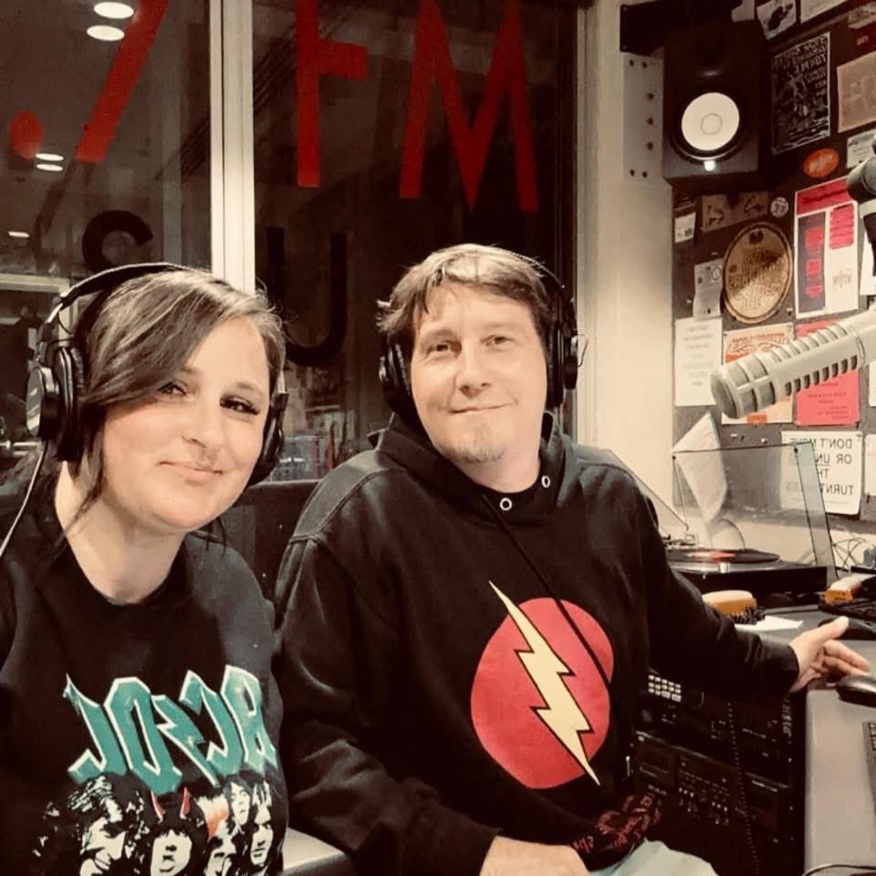 Two smiling radio hosts wearing headphones pose together inside a JFM radio station studio.