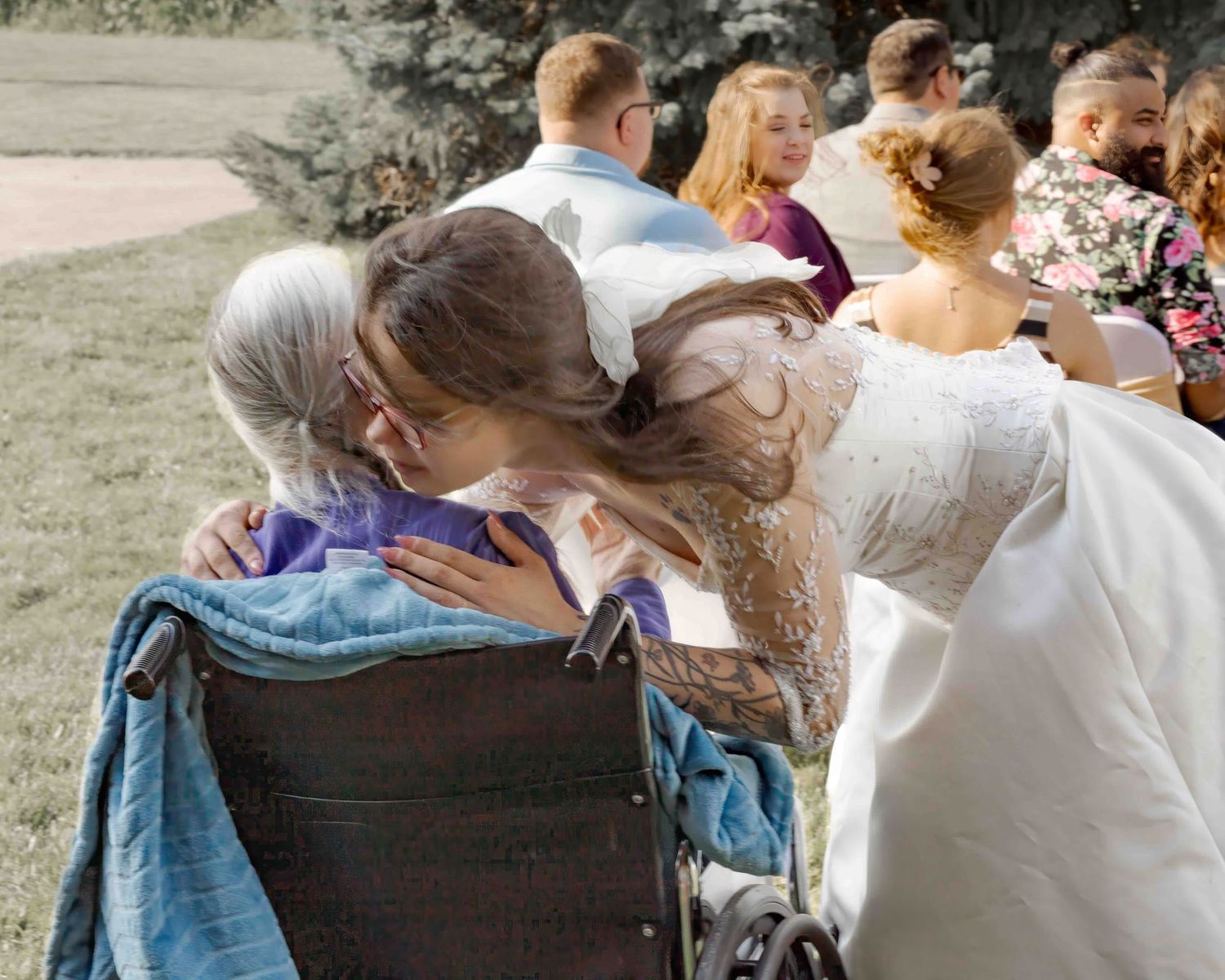 Bride in white lace gown embracing an elderly woman in a wheelchair at an outdoor wedding reception.